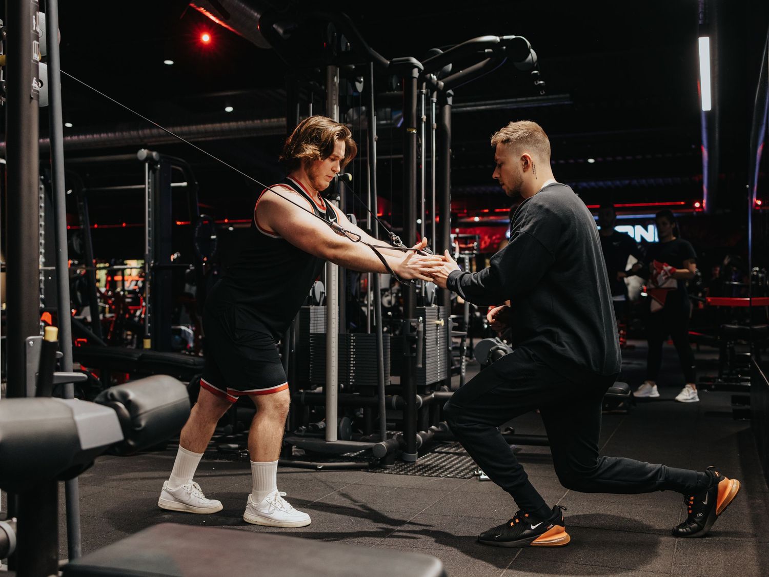 People working out in a dark modern gym with red lighting doing squats and barbell exercises.