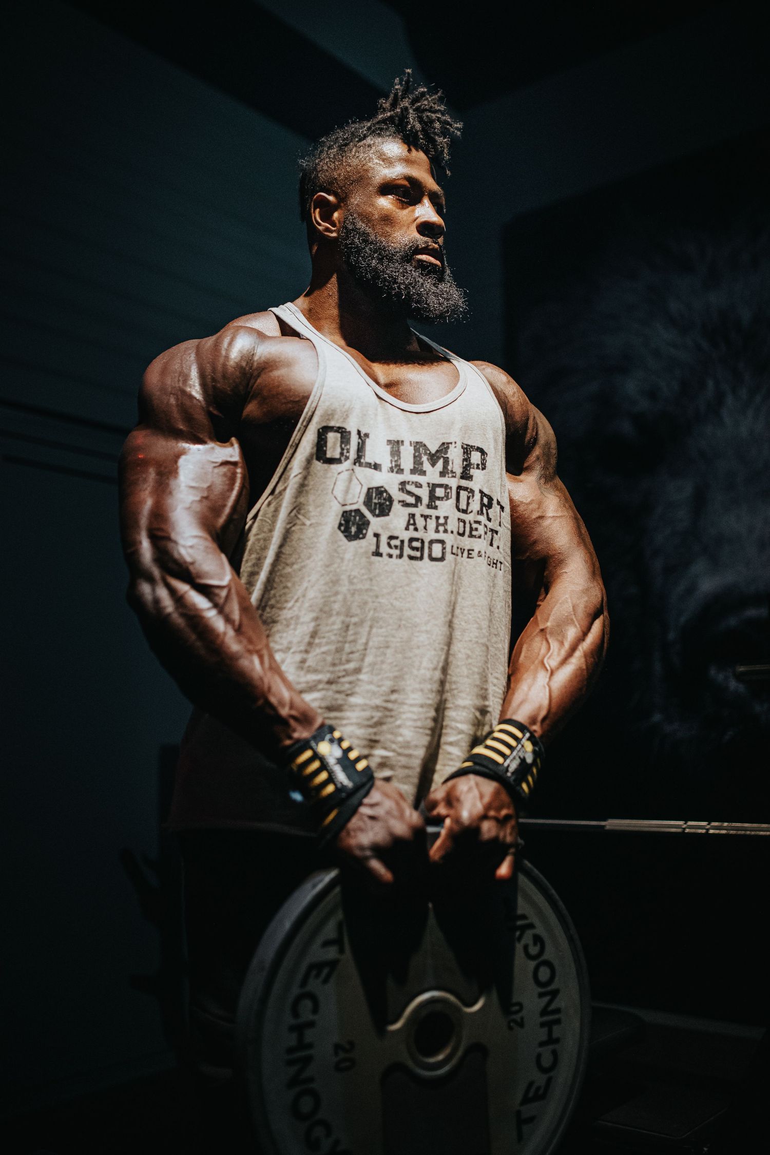 Muscular athlete in Olimp tank top holding barbell weight plate in dramatic dark gym lighting.