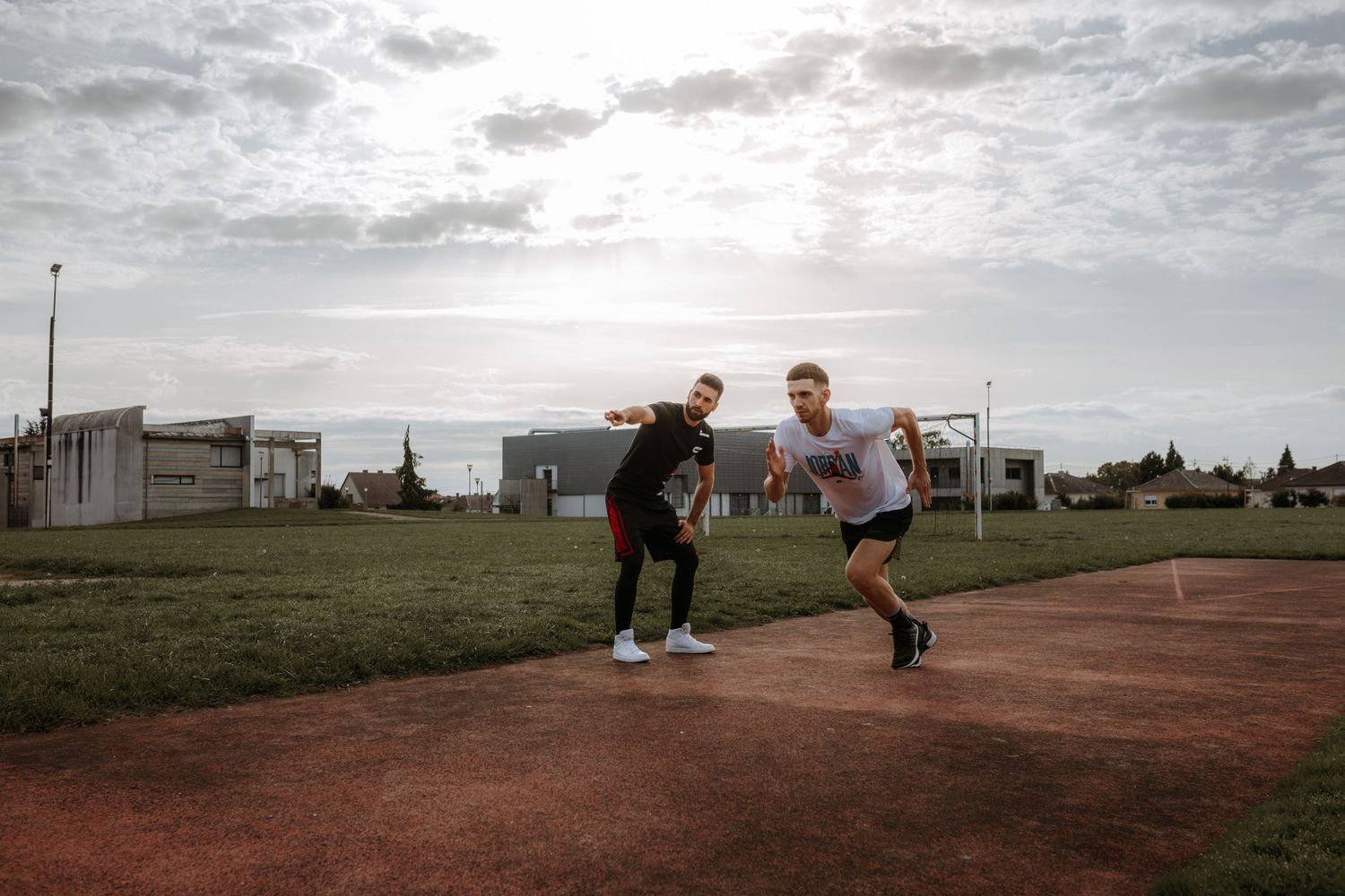 Two athletes train together at dusk on a dirt running track with houses and cloudy sky in the background.