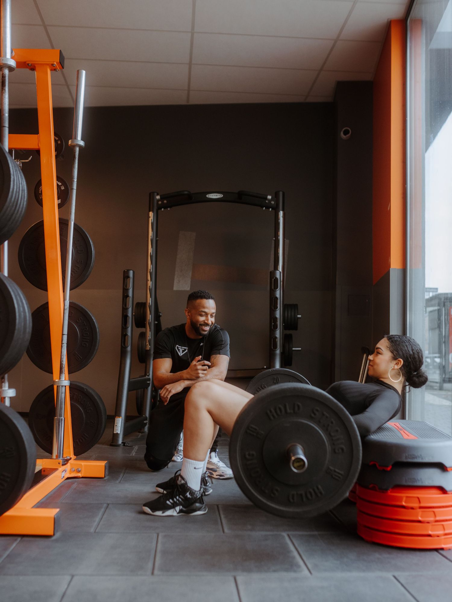A gym training area with weightlifting equipment including weight plates, orange storage racks, and mats along the wall.
