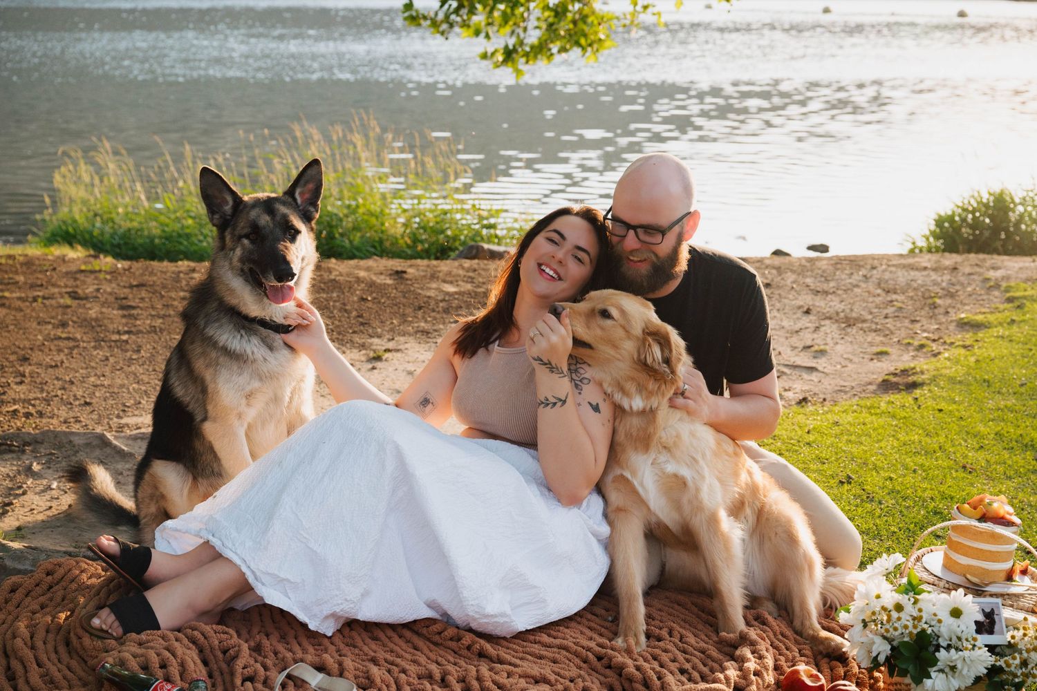 Two dogs joining a couple enjoying a lakeside picnic on a blanket during a sunny afternoon.