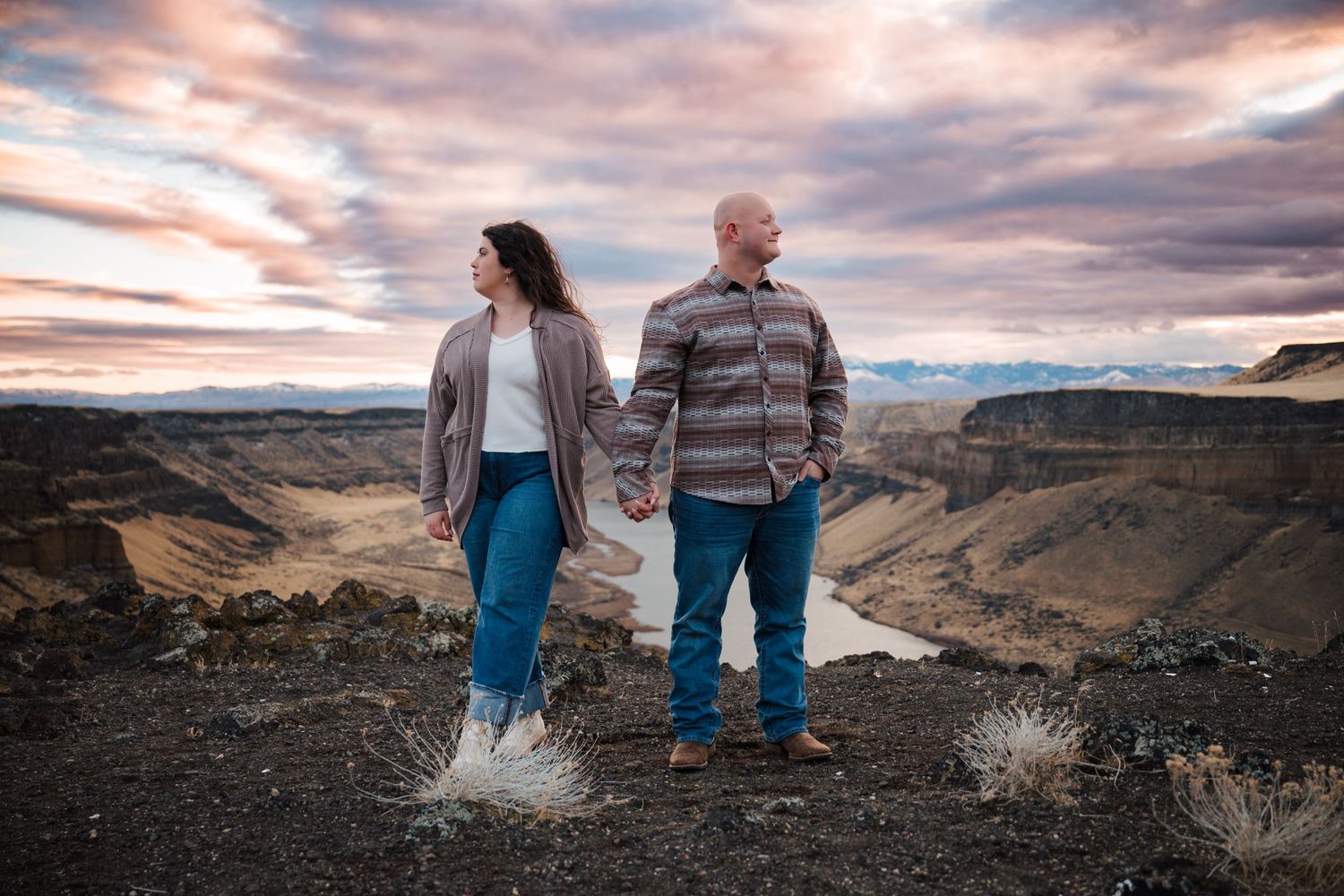 A couple holds hands while standing on a cliff edge overlooking a dramatic river canyon landscape.