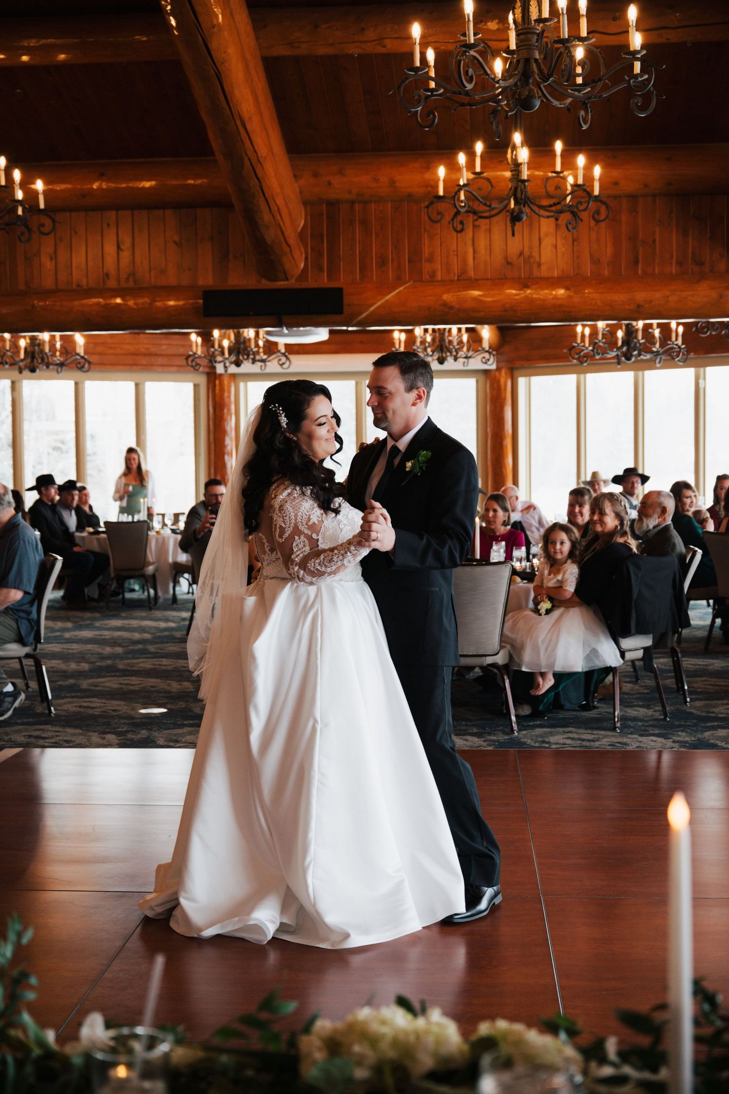 Wedding couple dancing in a rustic wooden venue with chandeliers and guests watching.