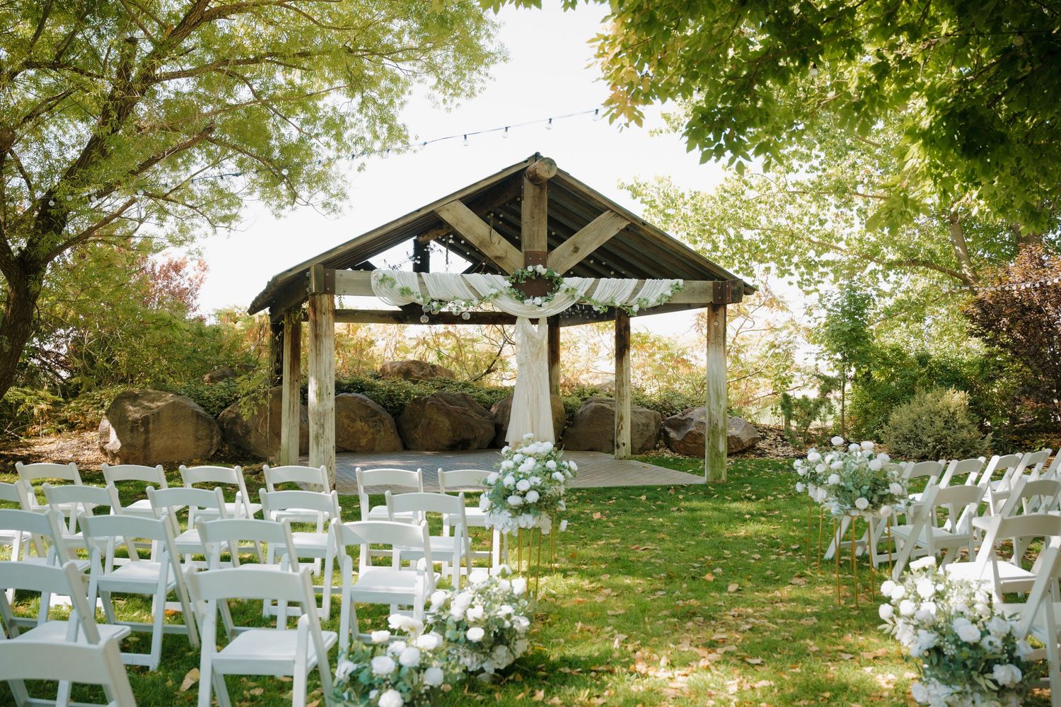 Rustic wooden pavilion with white stone columns set up for an outdoor wedding ceremony with white chairs on a lawn.