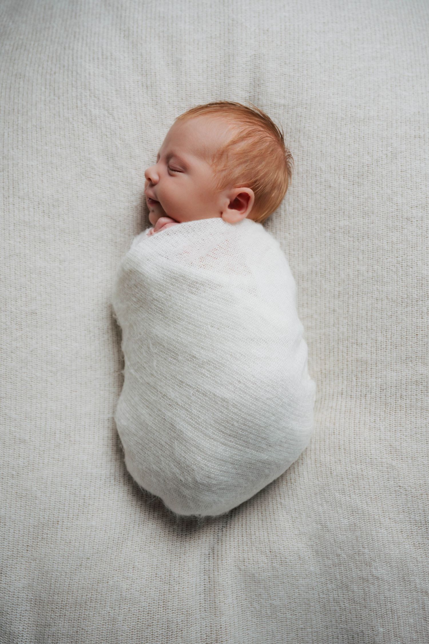 Newborn baby peacefully sleeping while swaddled in soft white muslin blanket on light gray background.