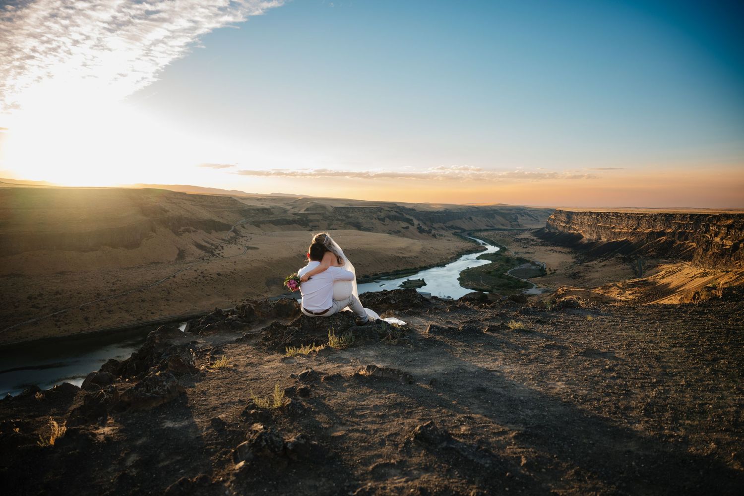 A couple wrapped in white fabric sitting on a cliff overlooking a winding river at sunset.