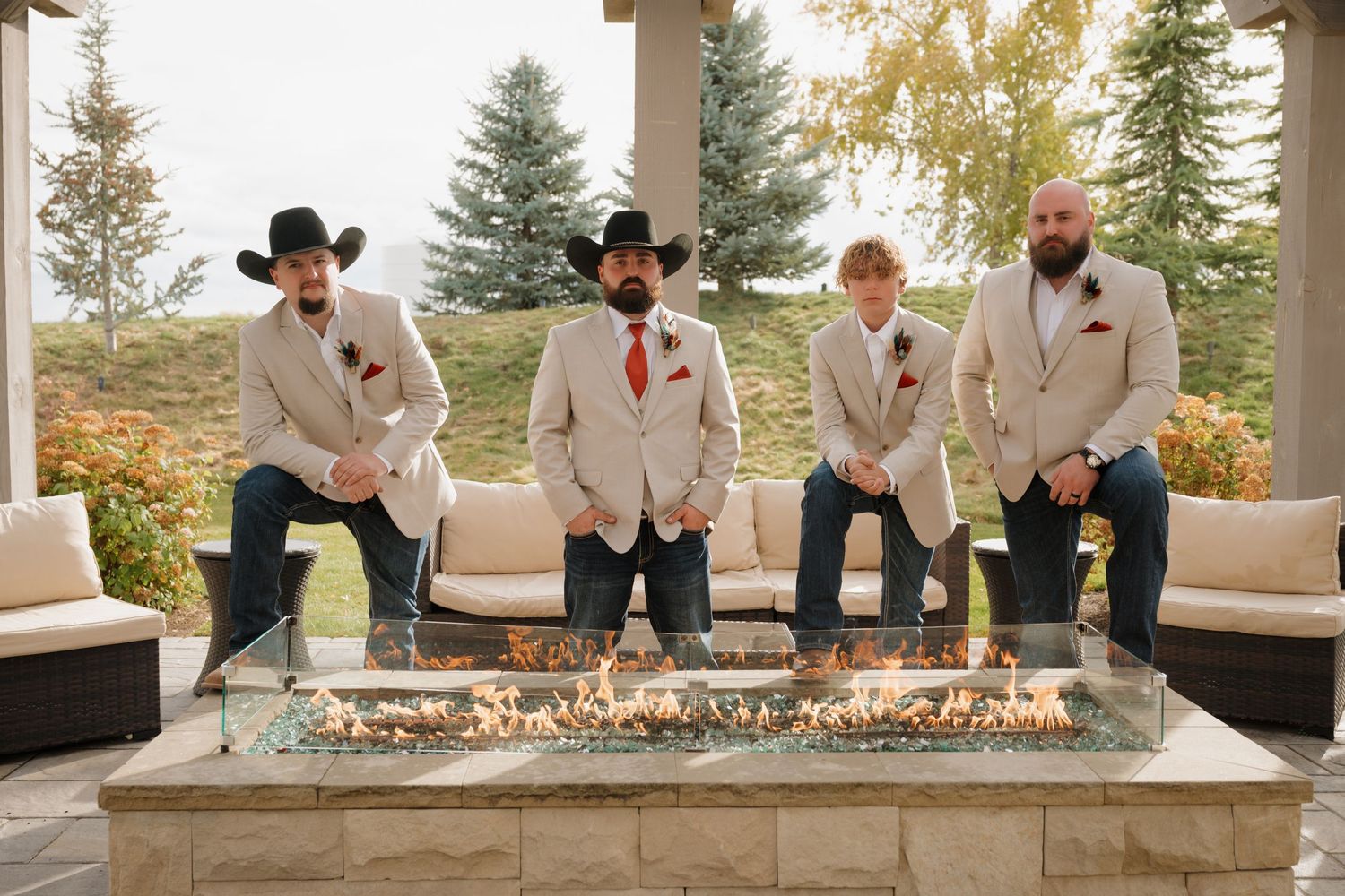Groomsmen in white jackets and cowboy hats gather around outdoor fire feature at sunset.
