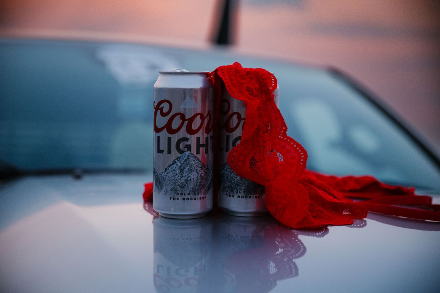 A can of Coors Light beer sits on a reflective surface next to vibrant red feathers at dusk.