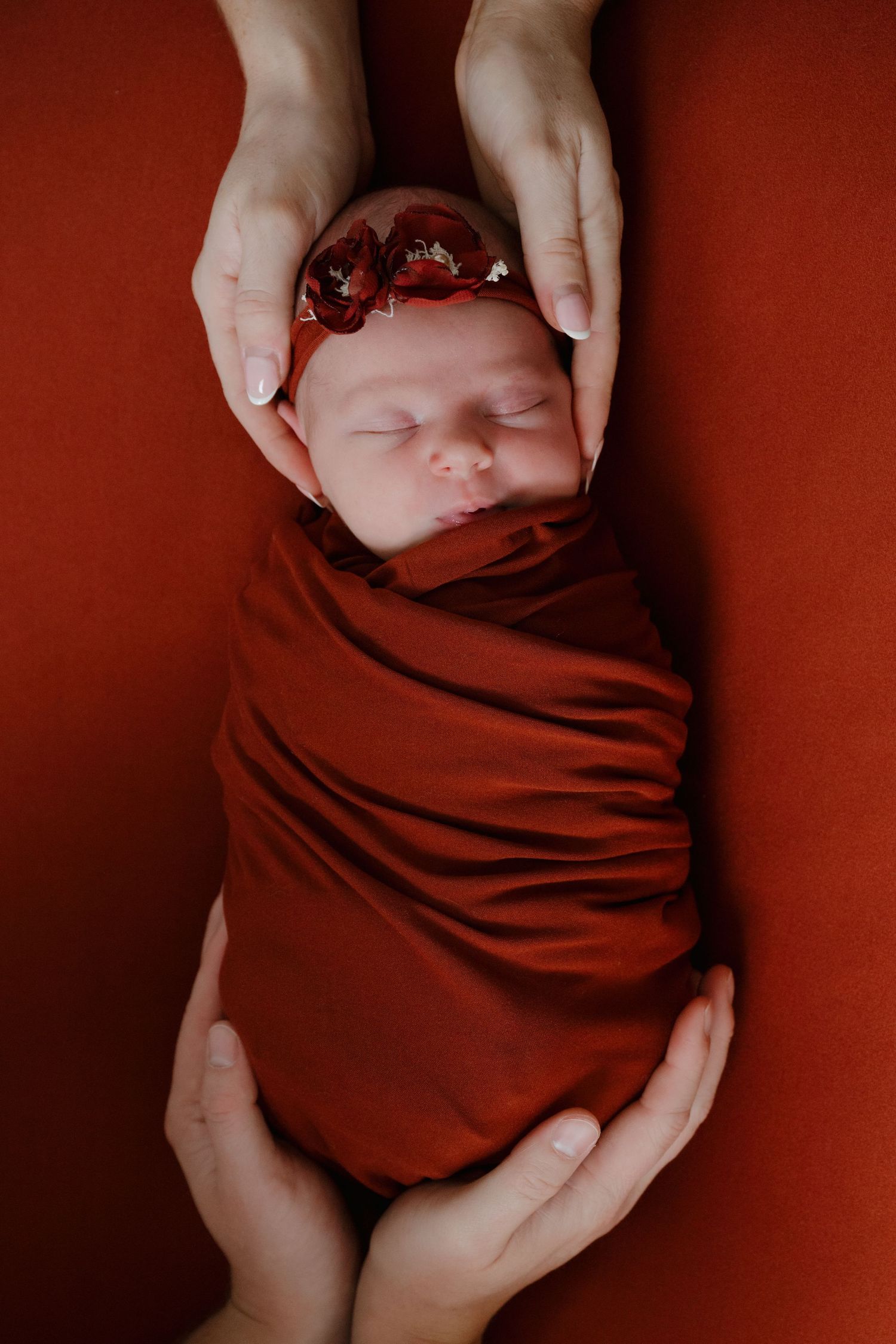 Sleeping newborn wrapped in deep red fabric with hands gently cradling their head.