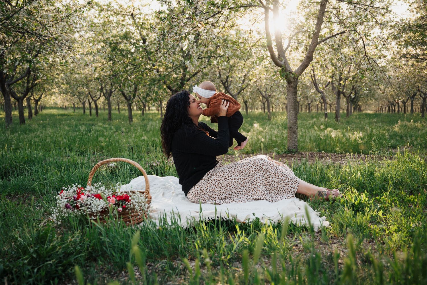 A picnic blanket with basket and flowers spread out in a serene spring orchard with sunlight filtering through trees.