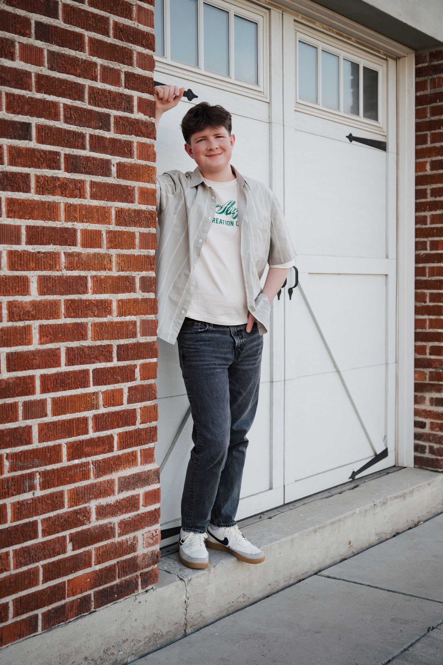 A person wearing a gray jacket and jeans poses casually against a brick wall next to white garage doors.