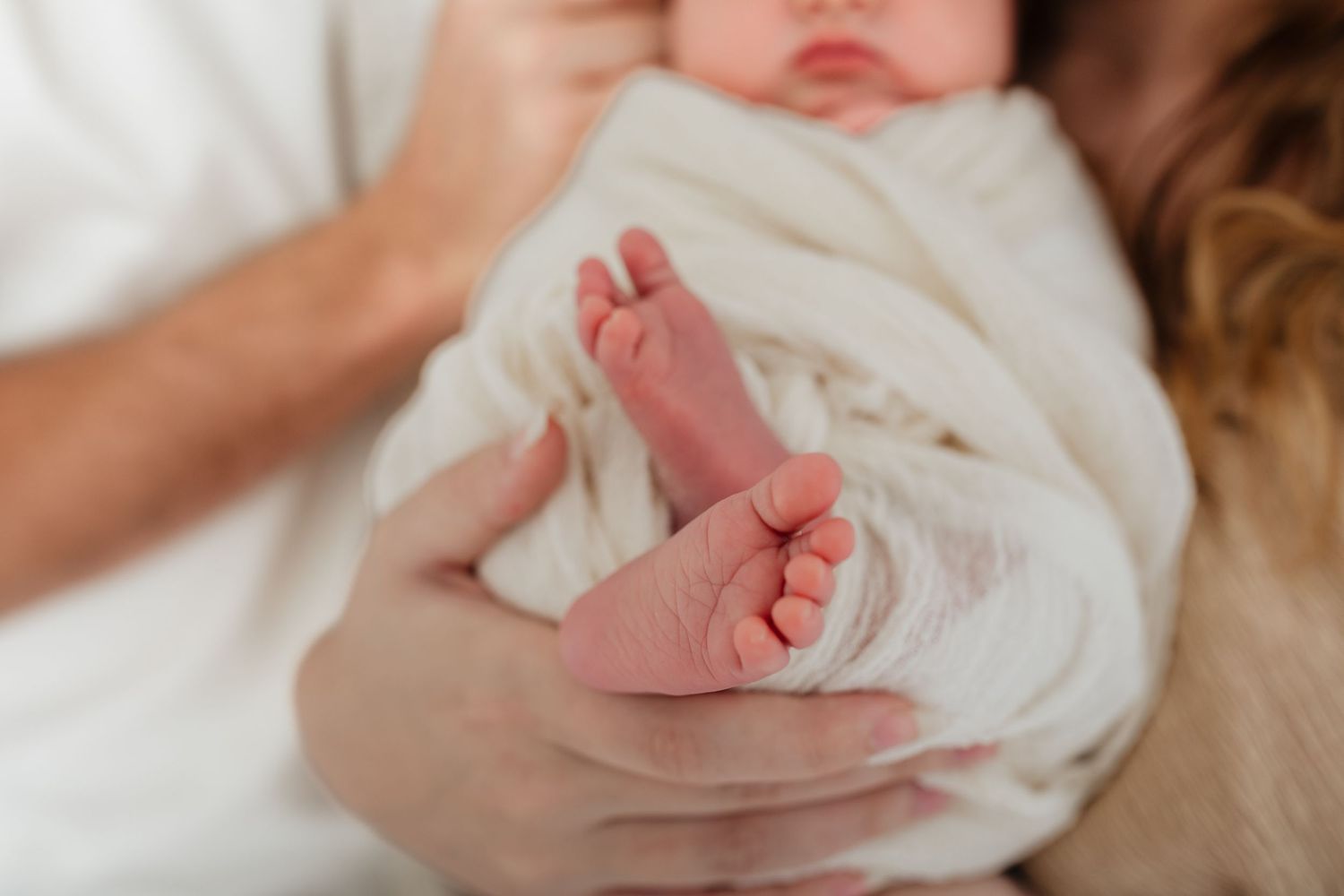Close-up of tiny newborn feet wrapped in white blanket with gentle hands holding.