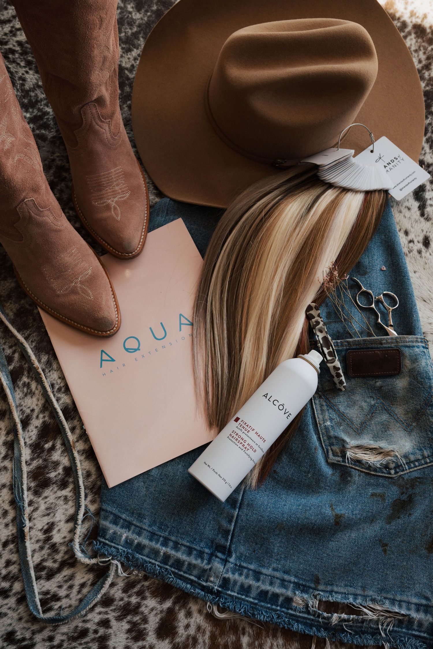 Flatlay of western boots, brown fedora hat, denim jeans, pink AQUA book, and beauty products on textured surface.