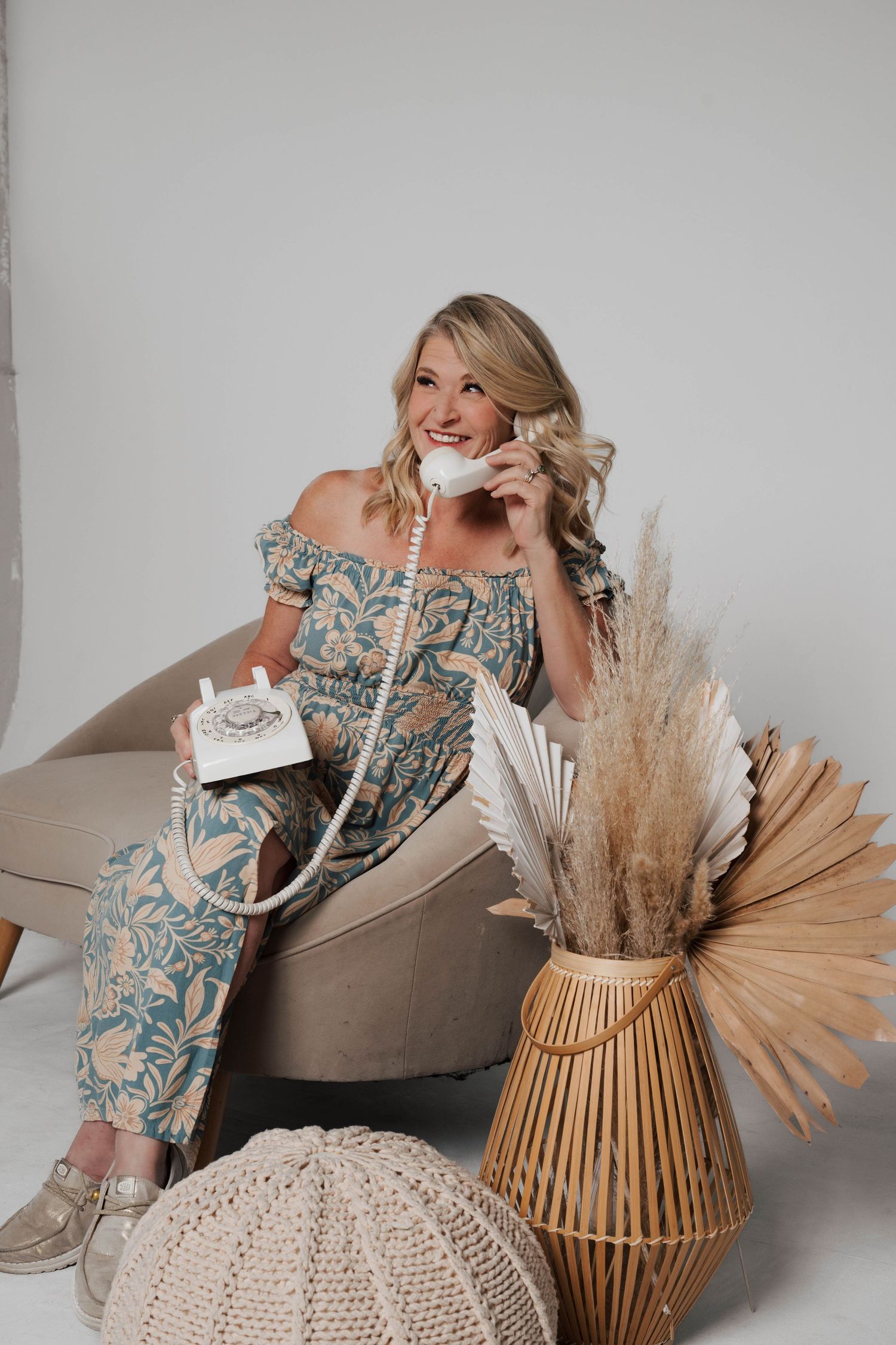 Woman in floral off-shoulder dress poses with vintage phone on couch surrounded by boho decor and pampas grass.