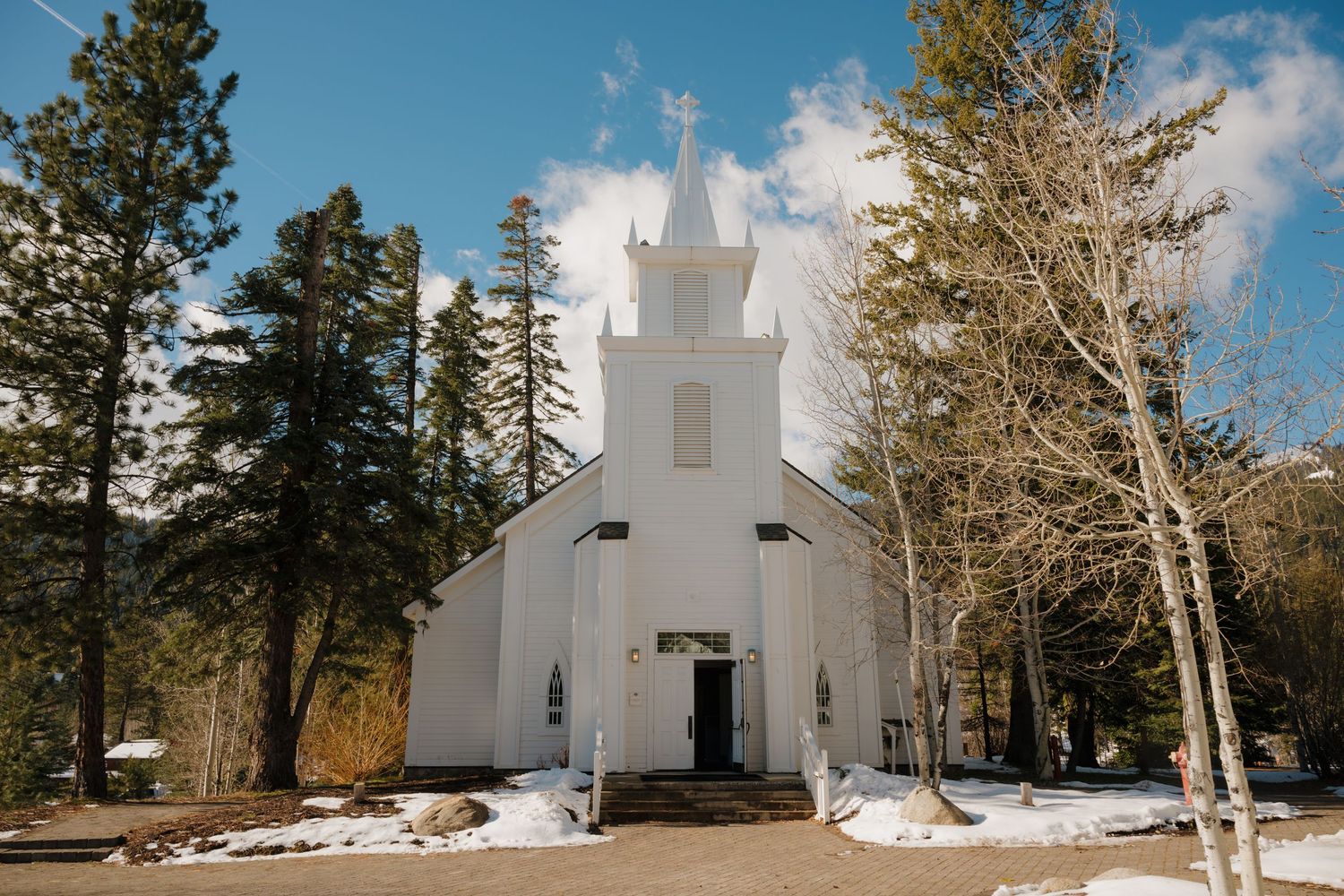 Snow surrounds a charming white wooden church with a steeple nestled among tall pine trees on a sunny winter day.