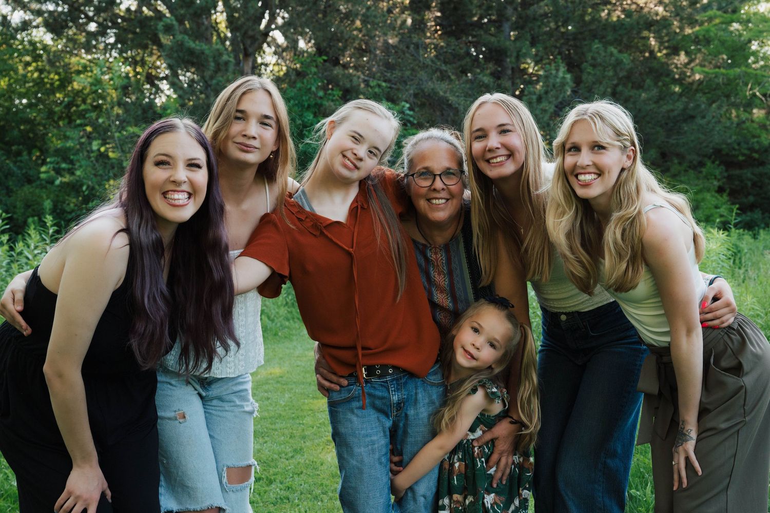 A cheerful group of seven people pose together outdoors on a sunny day with green trees in the background.
