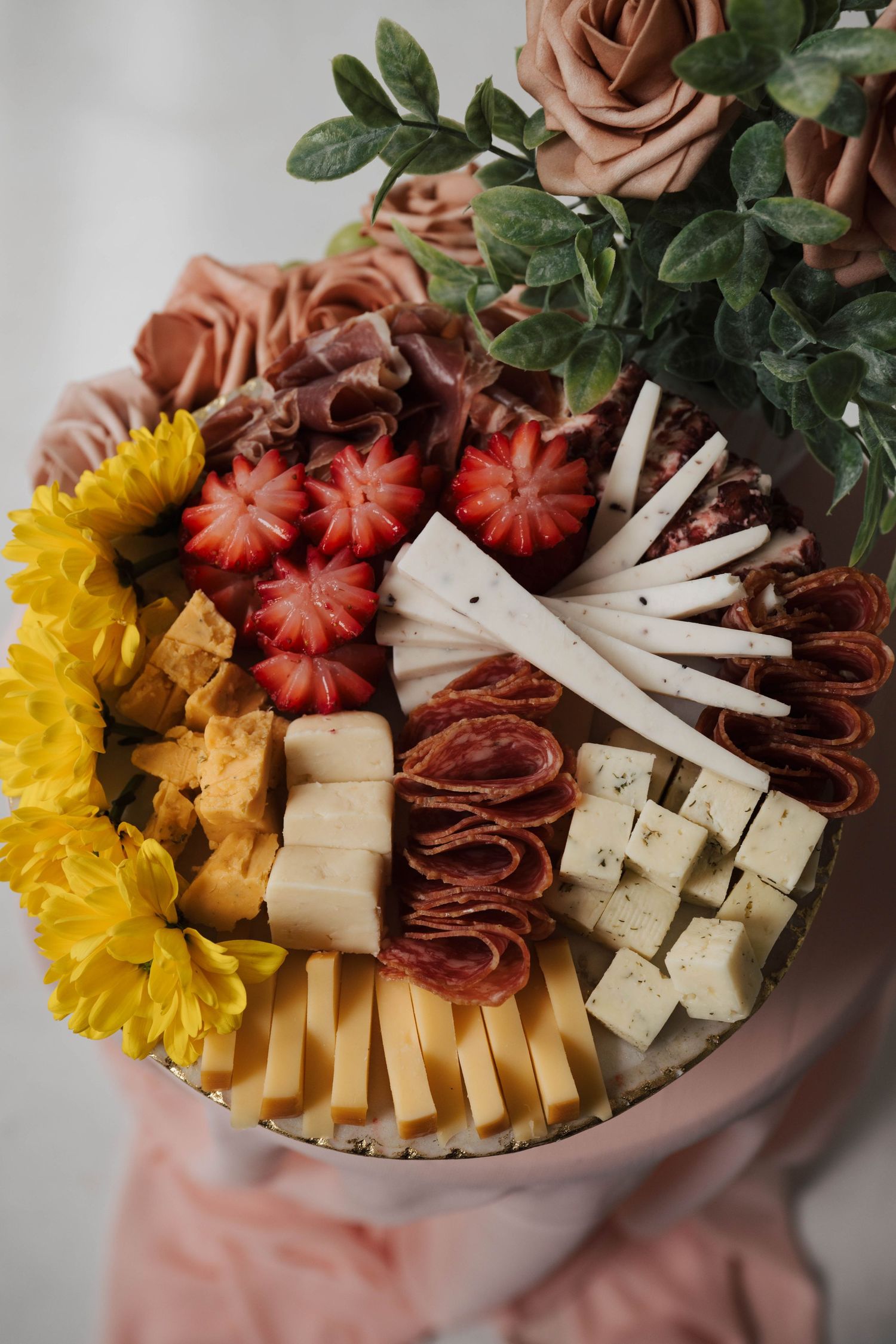 Charcuterie board featuring salami, cheese, strawberries and yellow flowers arranged with dried roses in the background.