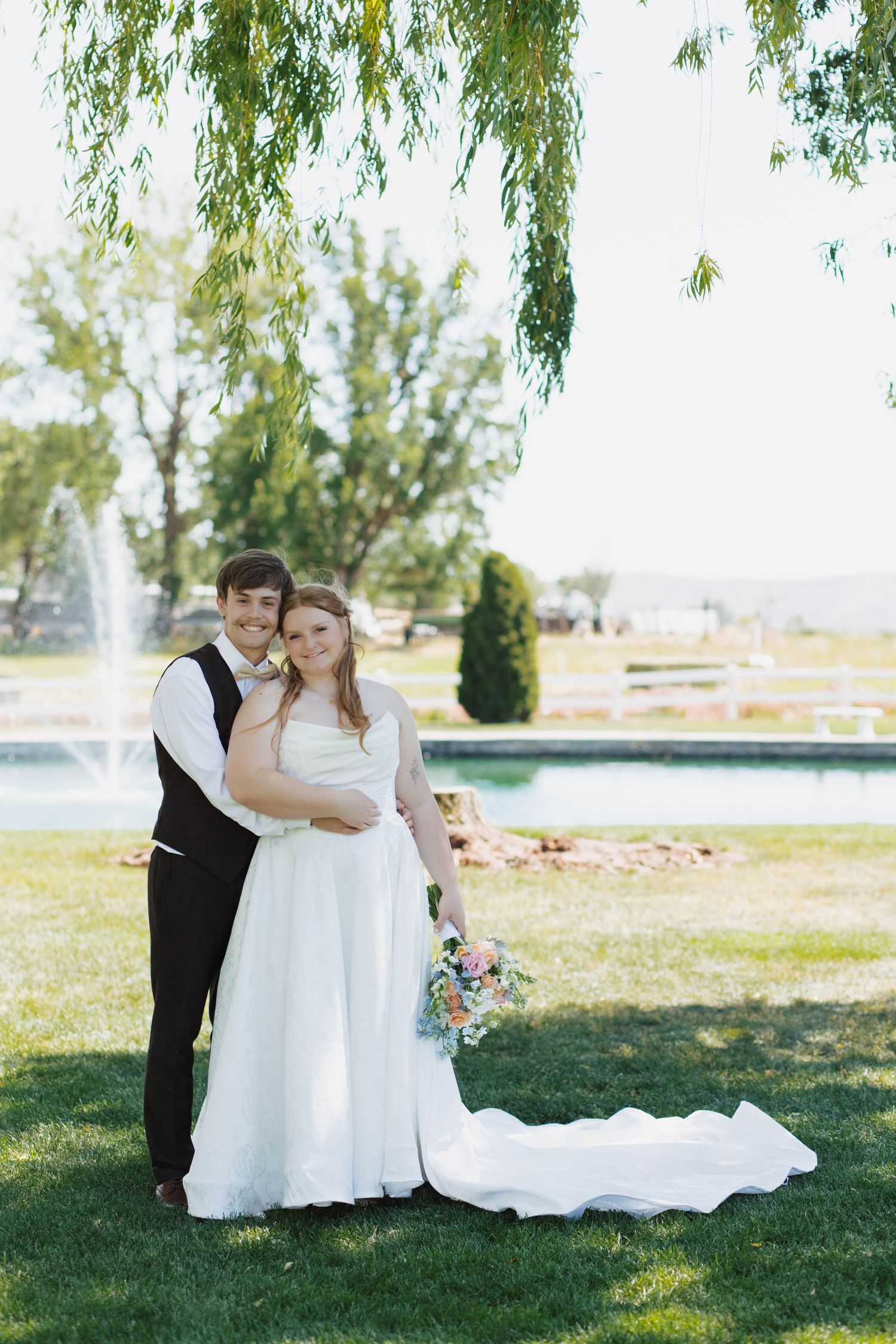 A couple stands together on a manicured lawn near a fountain, with willow trees and water features in the background.