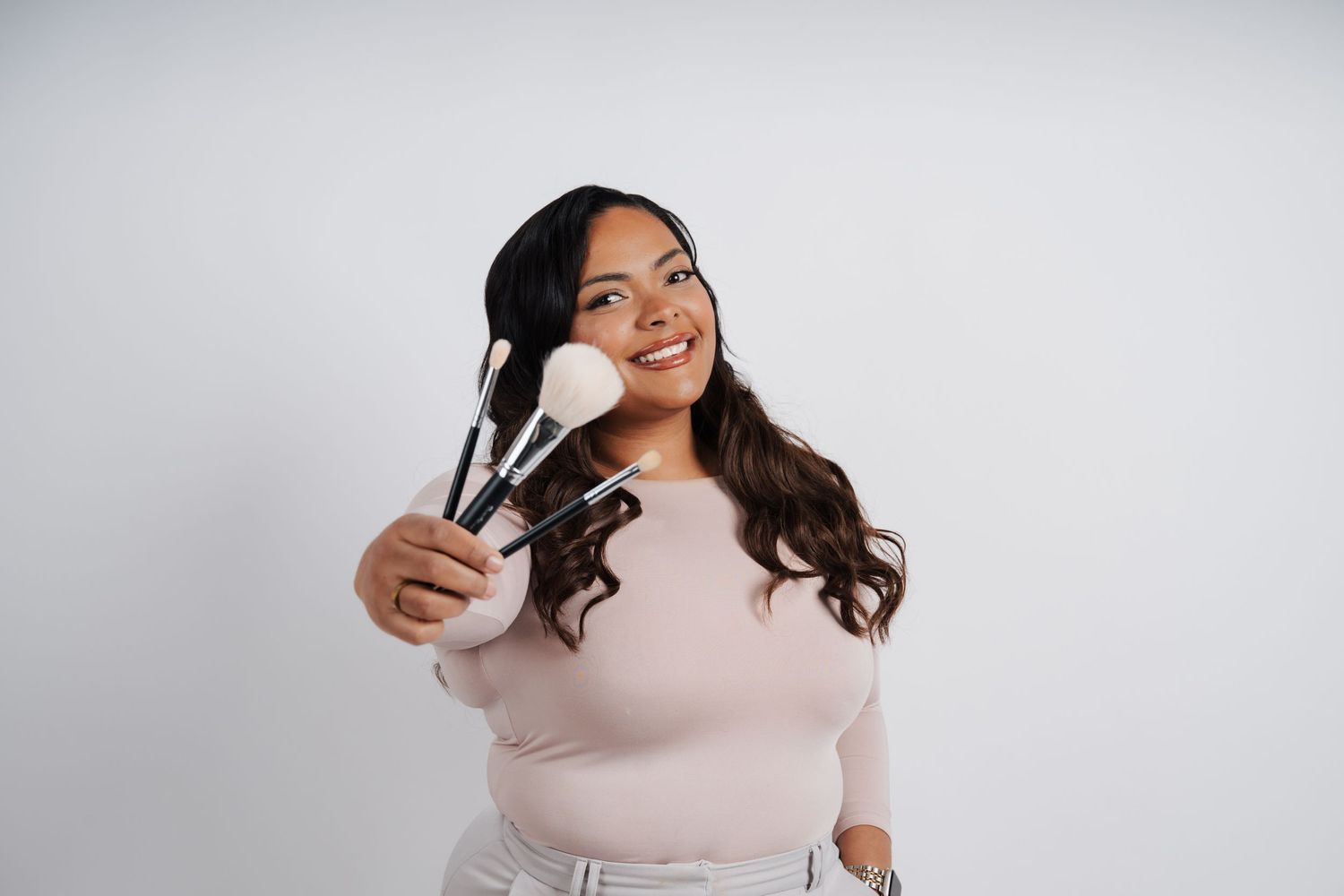 A smiling person wearing a cream sweater holds up makeup brushes against a light gray background.