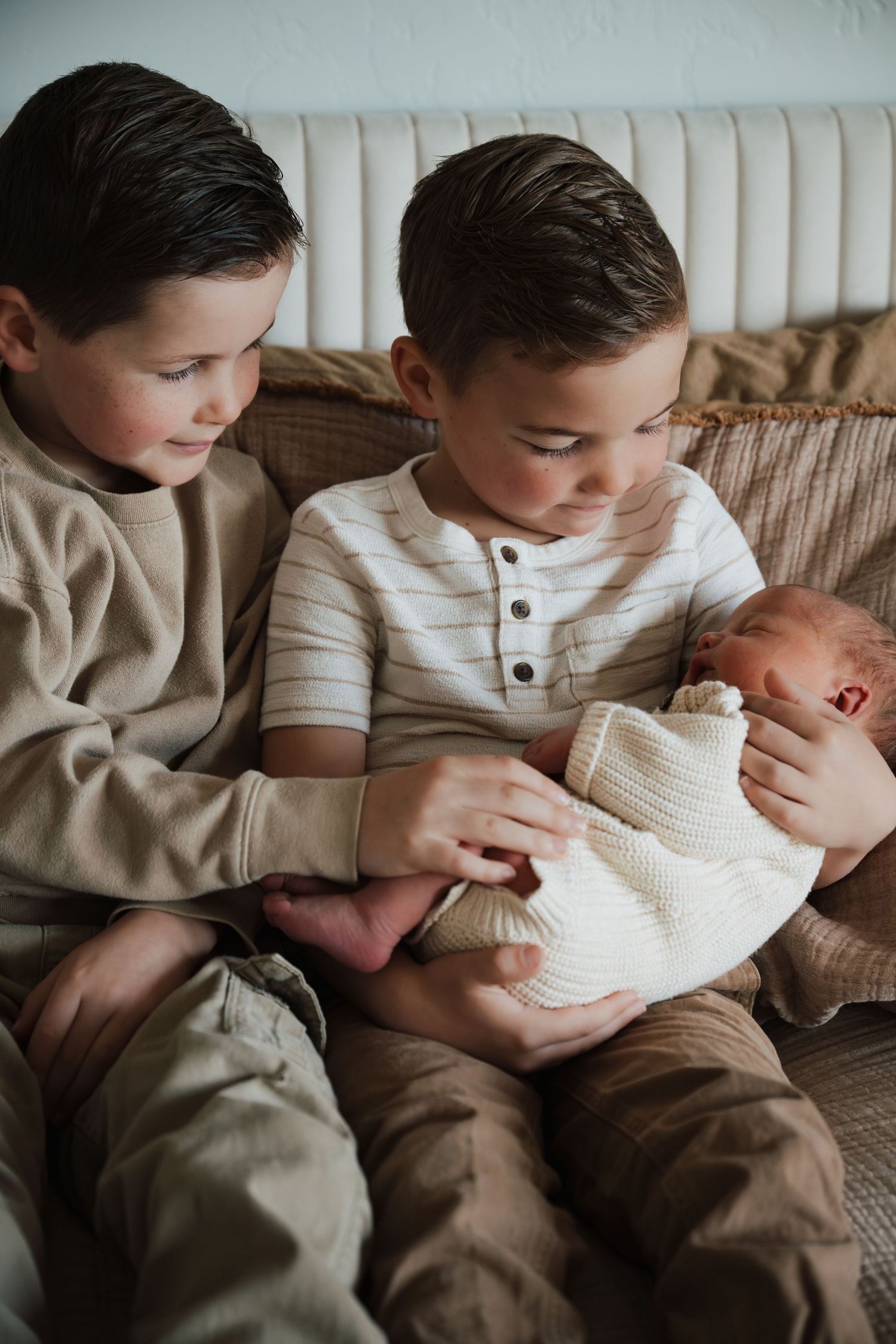 Two young boys sitting on a couch lovingly holding and looking at a newborn baby wrapped in a white knit blanket.