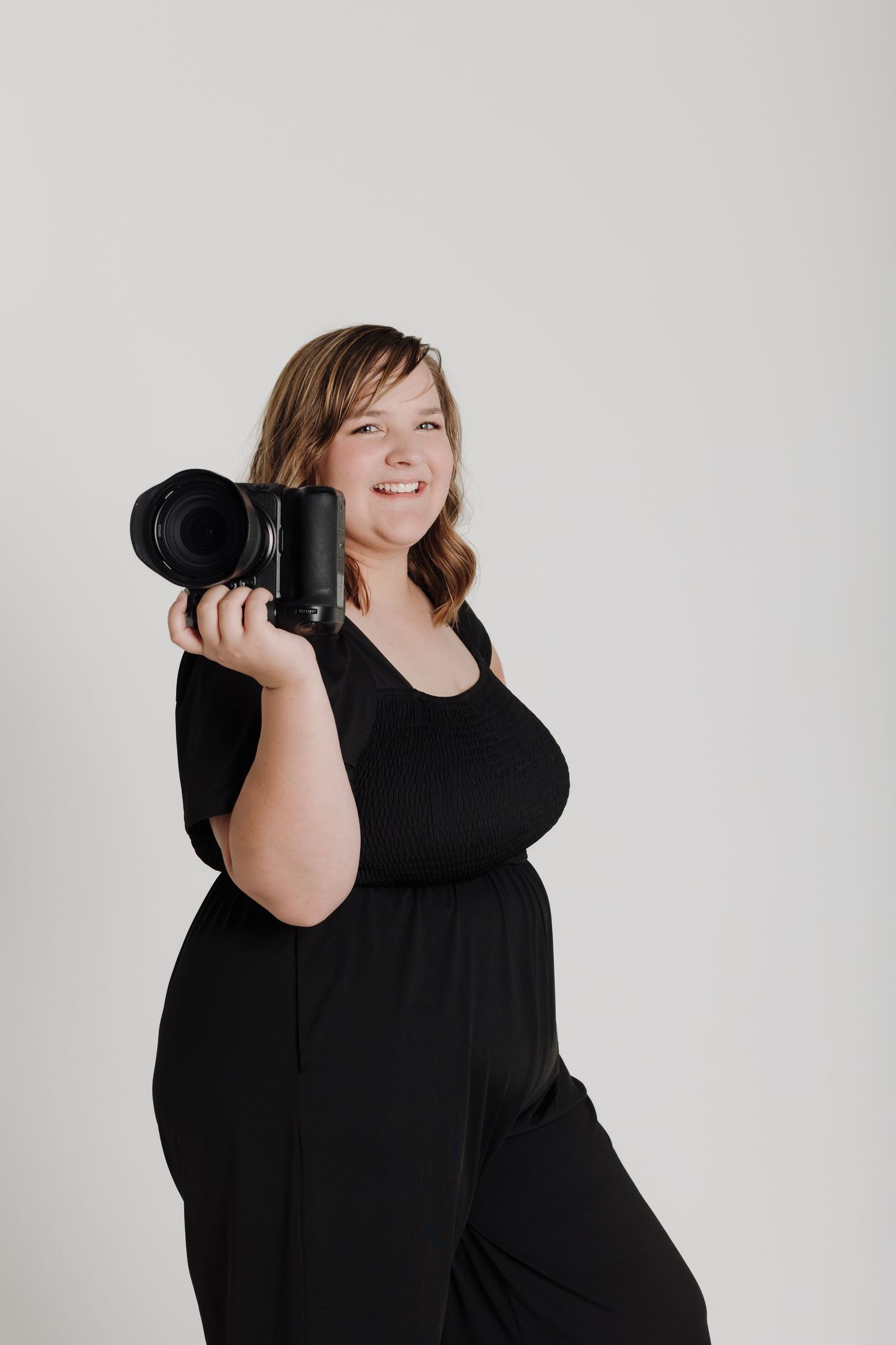 Portrait photographer in black dress holds professional camera while posing against studio background.