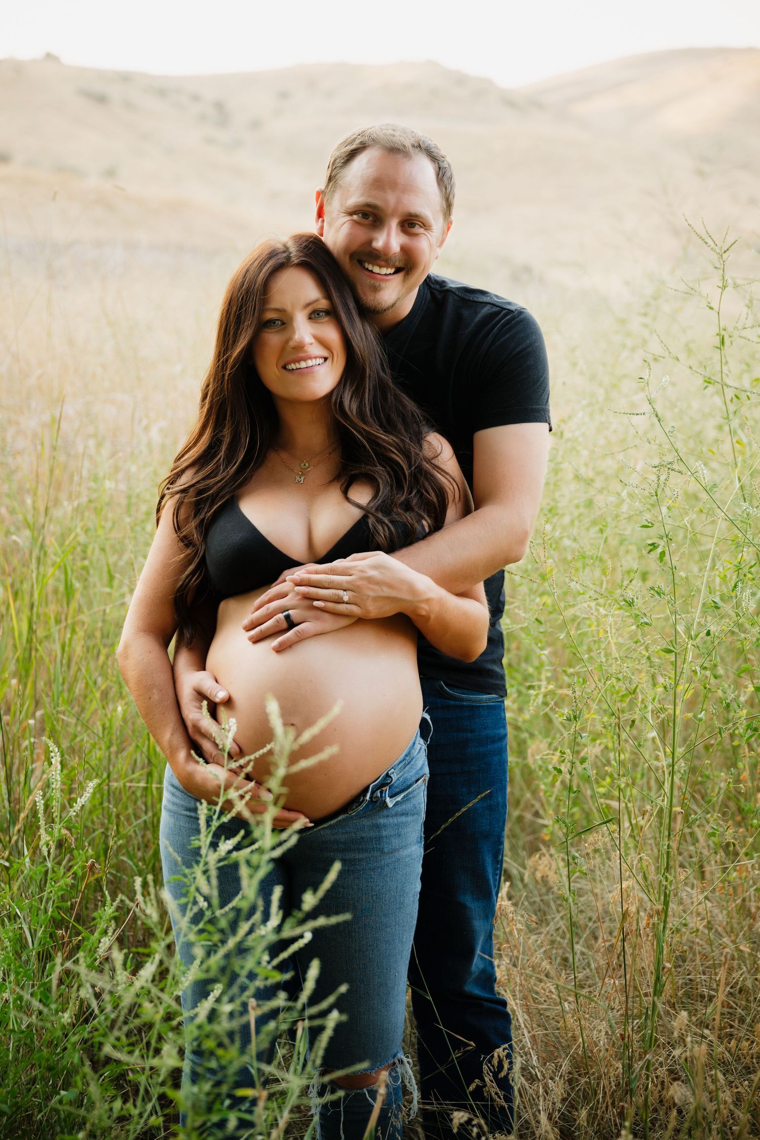 Expectant couple embraces in a sun-lit meadow with rolling hills in the background.