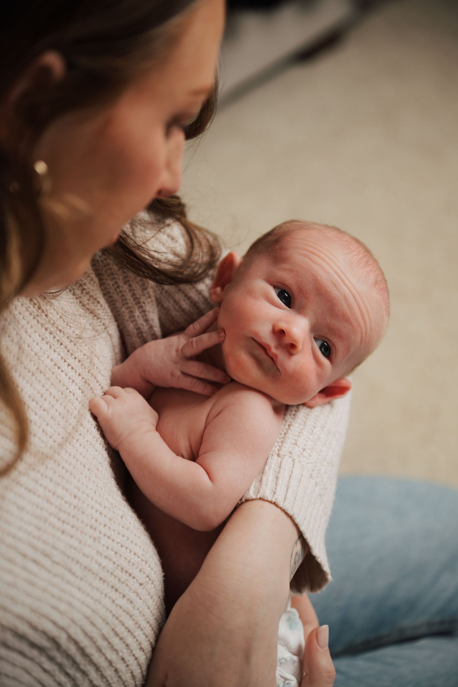 Parent in cream sweater holds alert newborn baby against their chest.