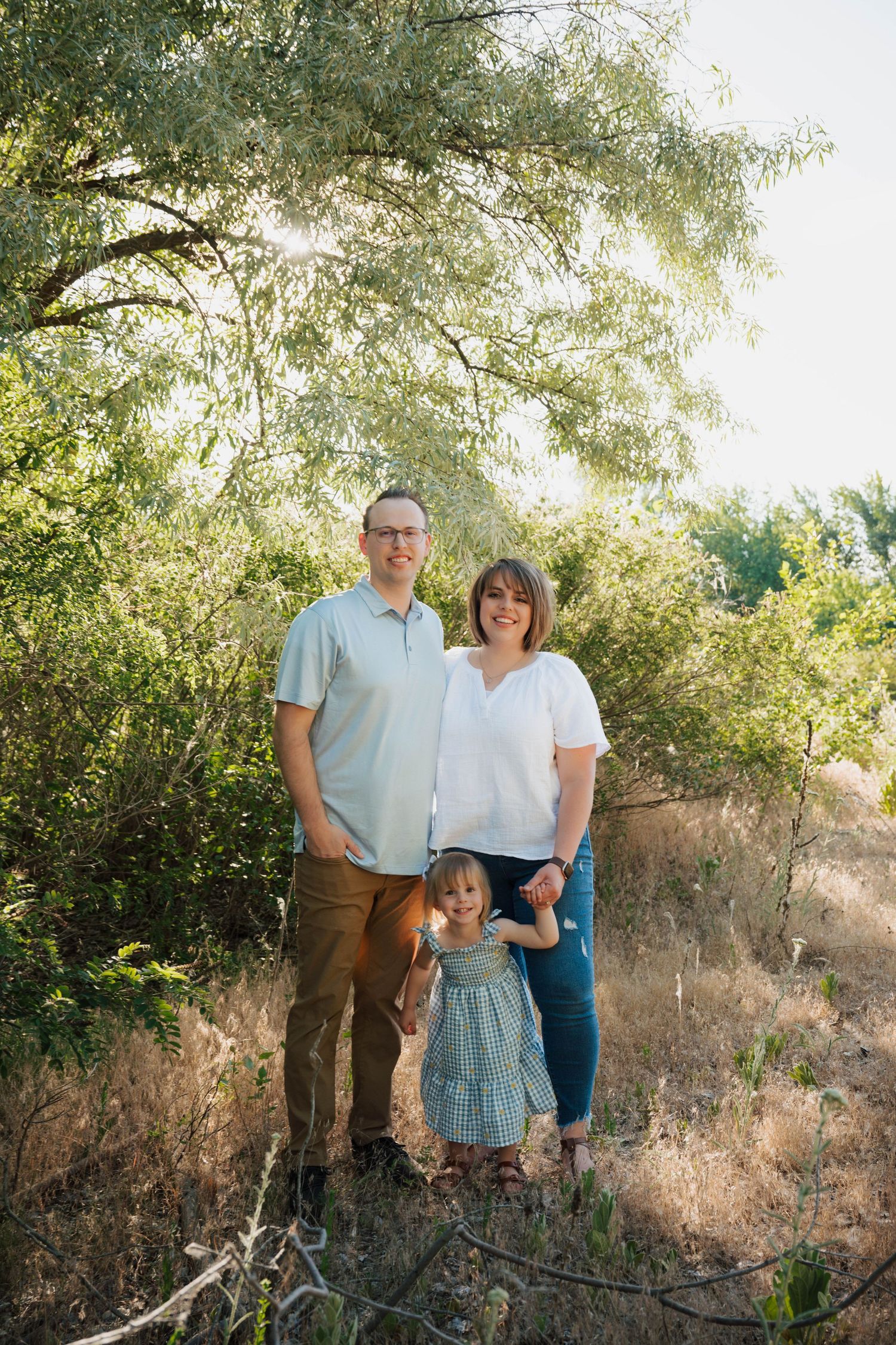 A family stands together outdoors against a backdrop of sunlit trees and dry grass in a natural setting.