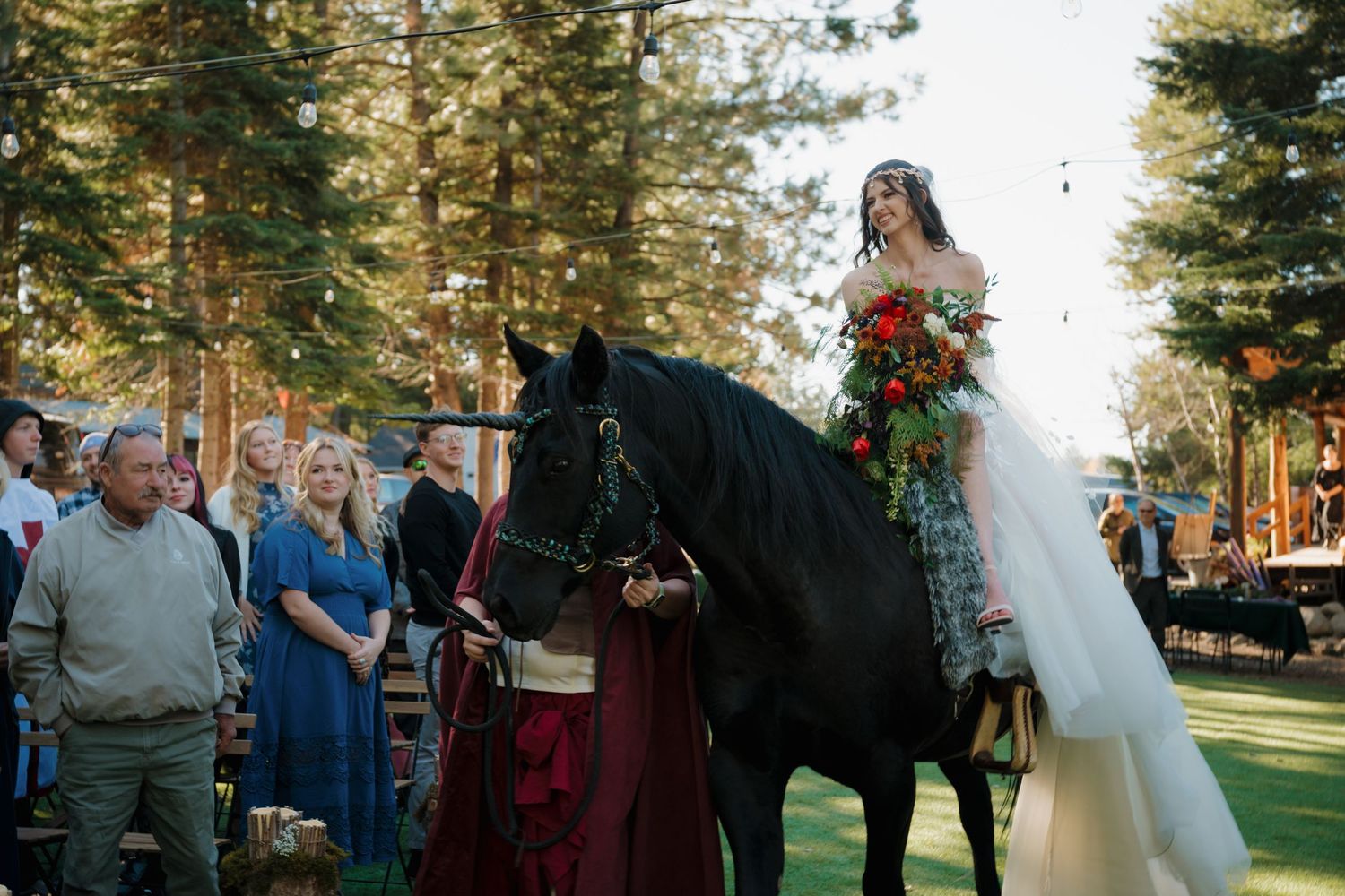 A bride on horseback wearing a white dress and holding red flowers during an outdoor wedding ceremony.
