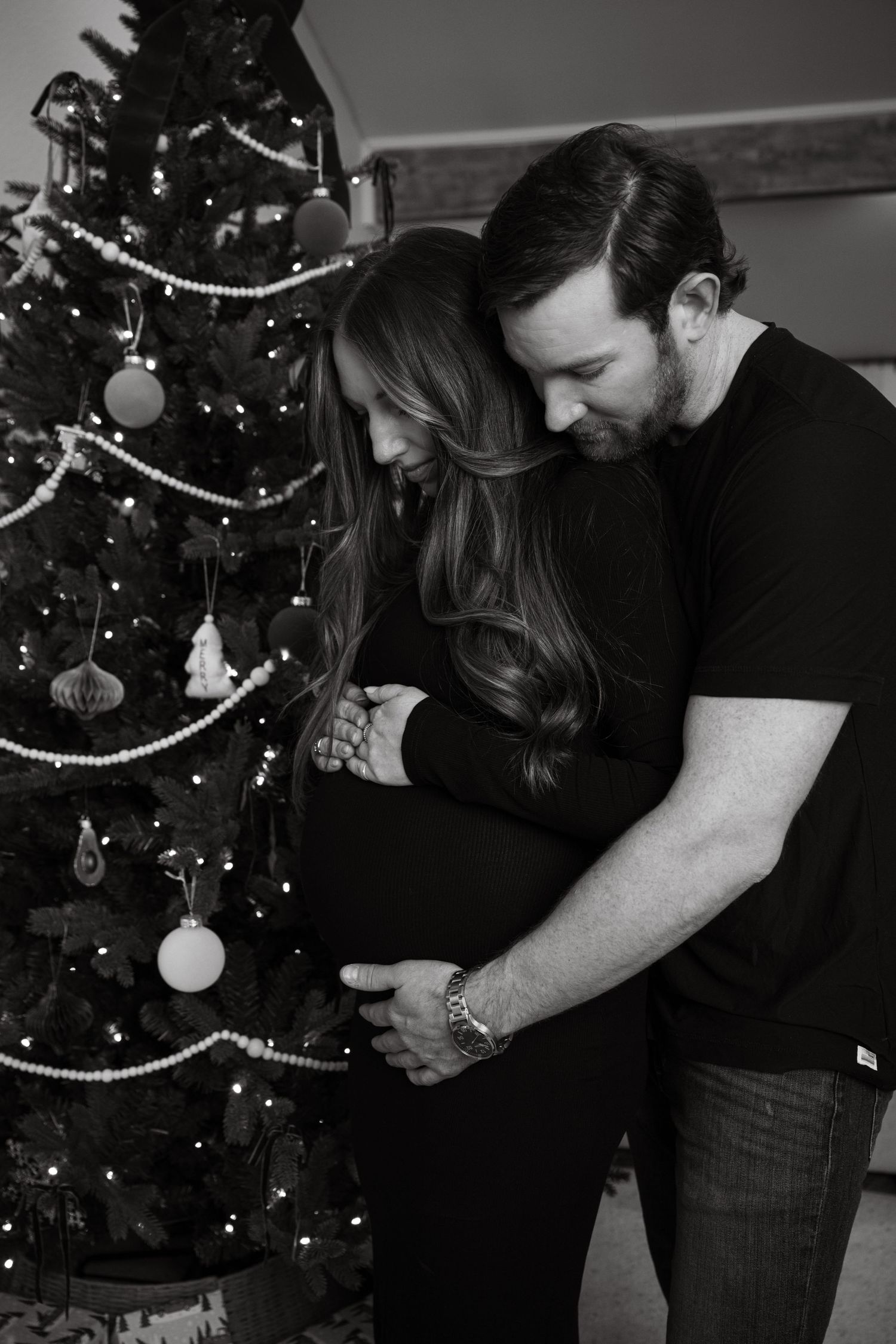 Black and white maternity photo of couple embracing by decorated Christmas tree.