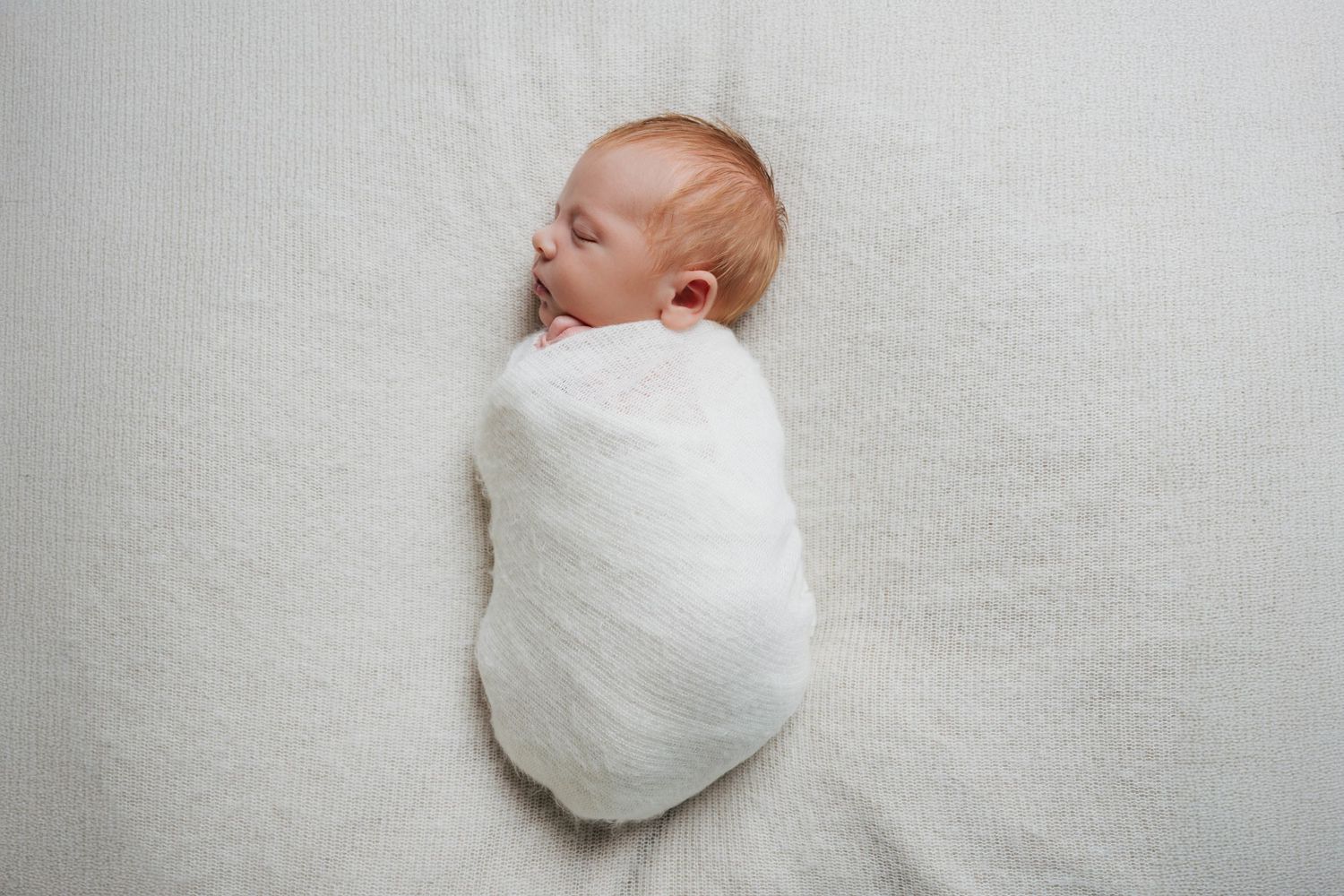 Sleeping newborn wrapped snugly in white swaddle blanket resting on cream textured surface.