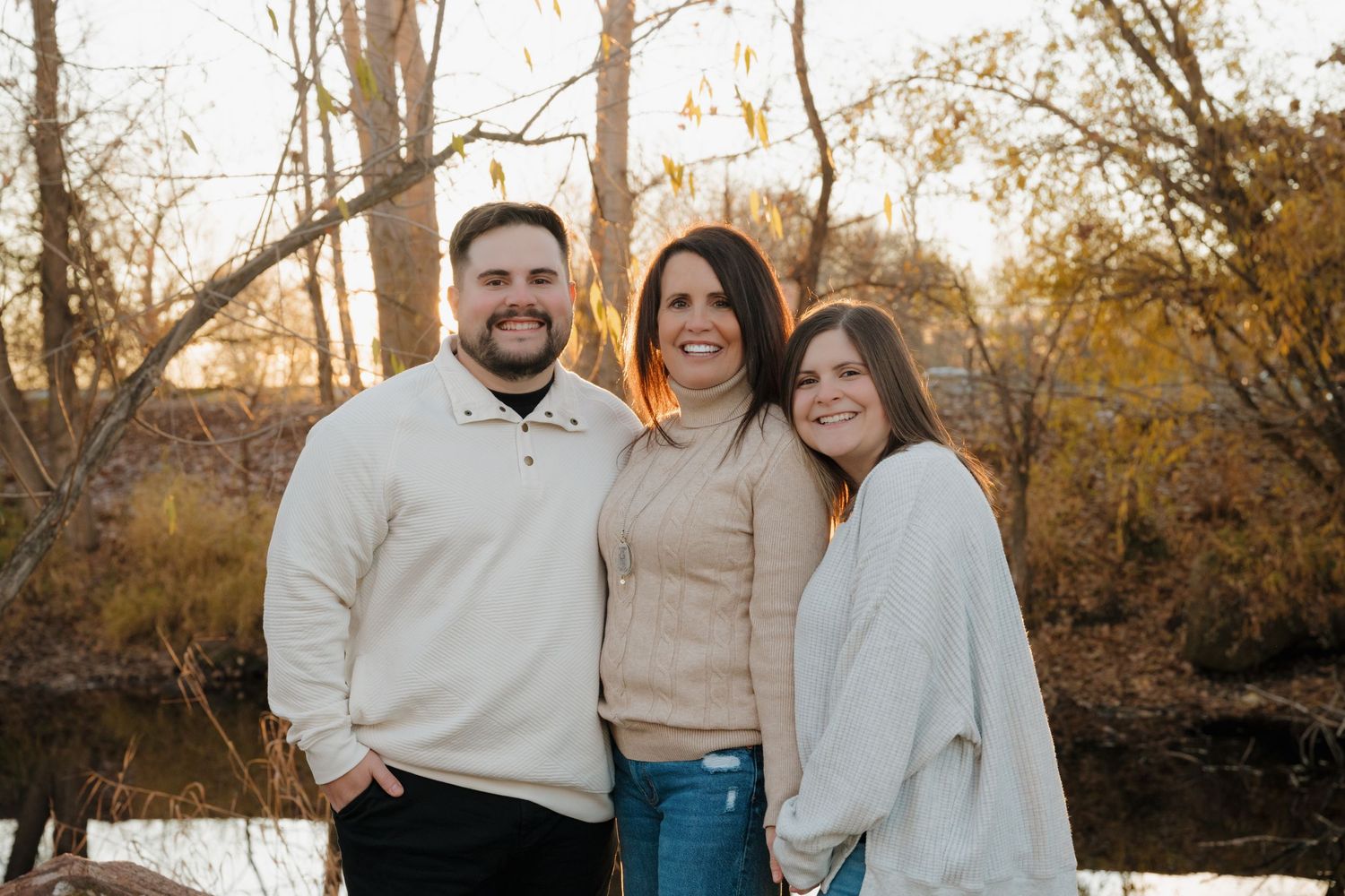 A family of three poses together outdoors against a scenic autumn backdrop with bare trees and a creek.