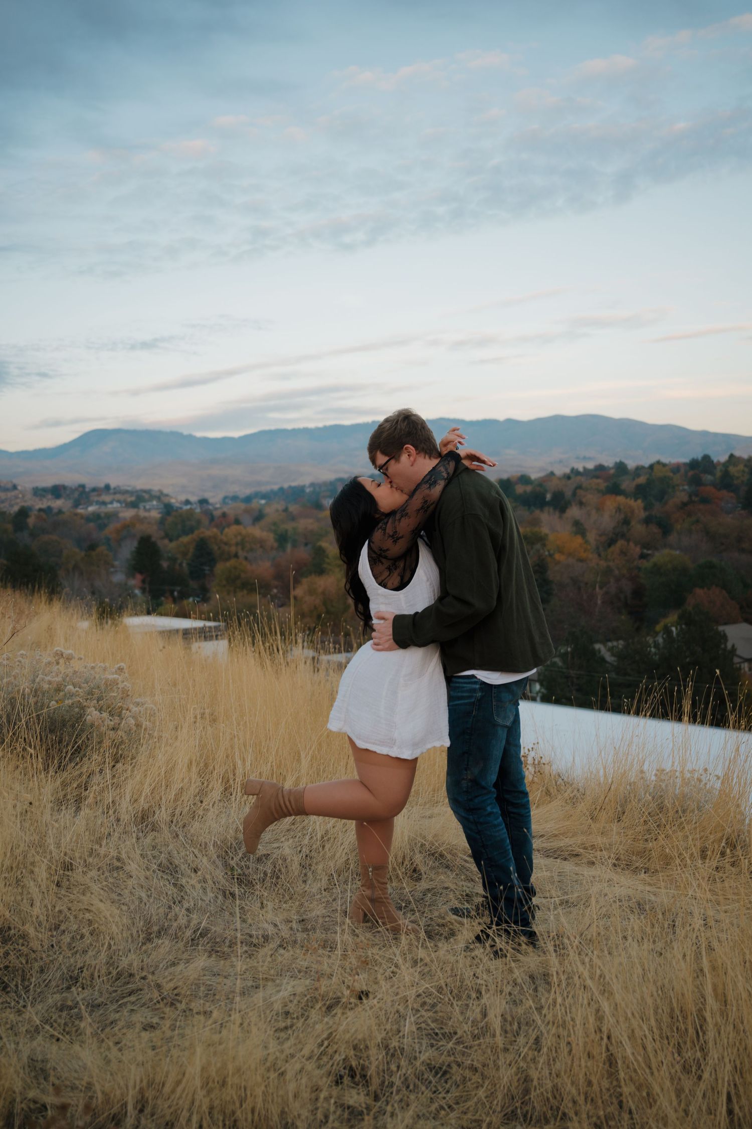 A romantic couple embraces in tall golden grass with mountains in the background at sunset.