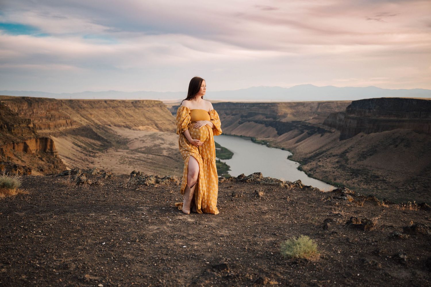 A long yellow dress contrasts against the dramatic landscape of a winding river canyon at sunset.