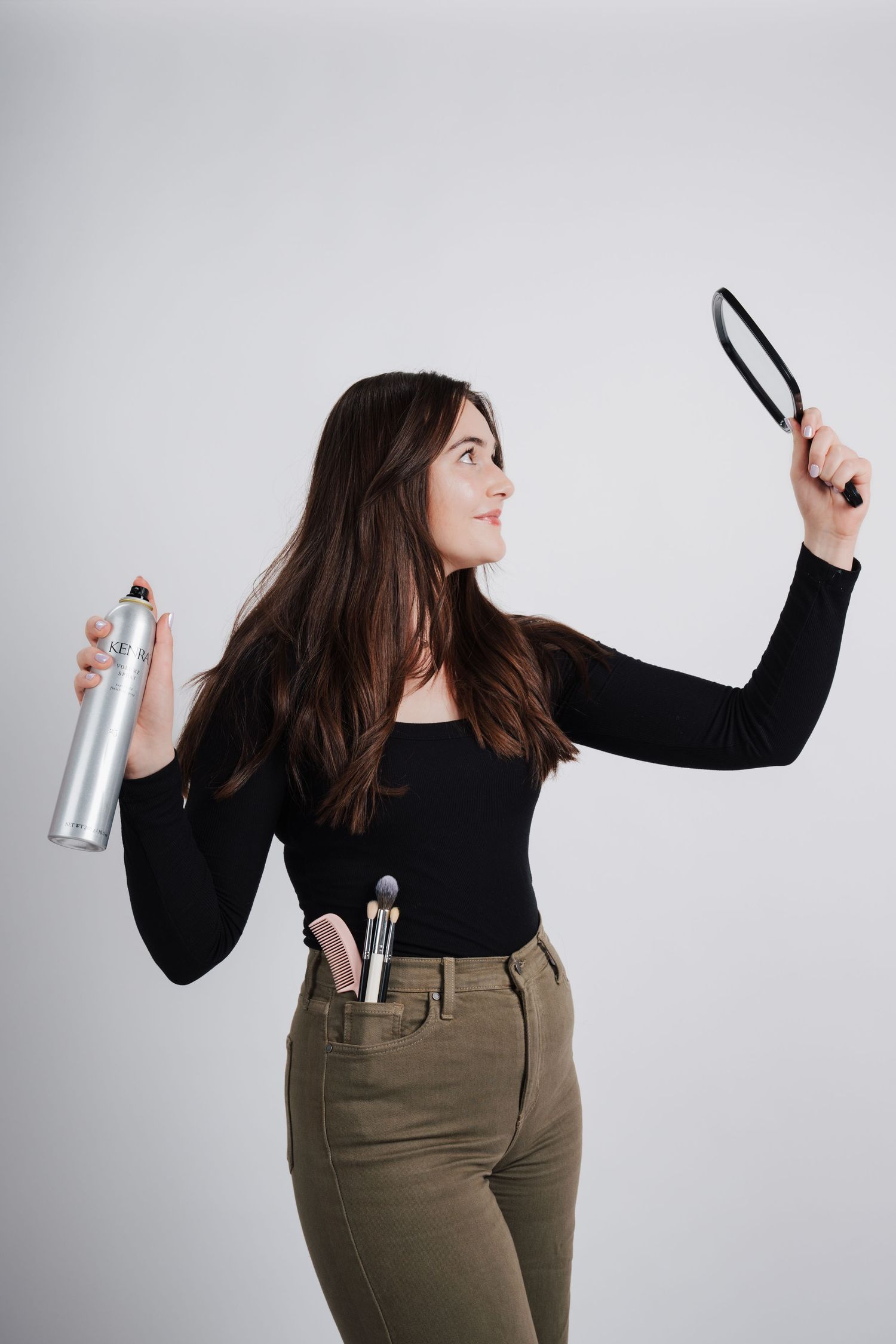 Woman in black long sleeve top and olive pants holds hair styling products against gray background.