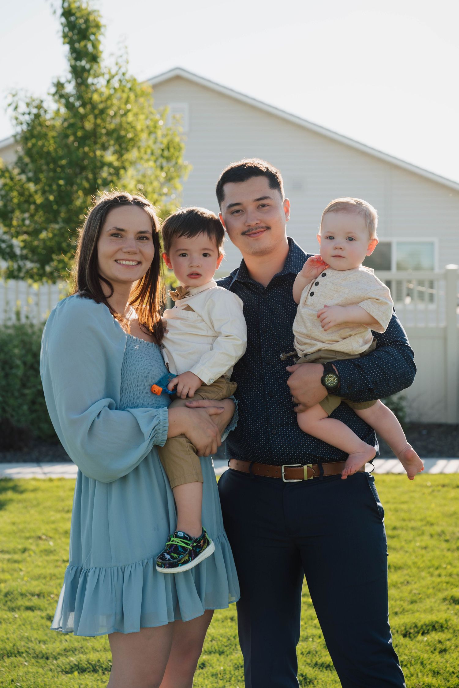 A family of four poses together on a sunny day in front of a white house with green grass and trees in the background.