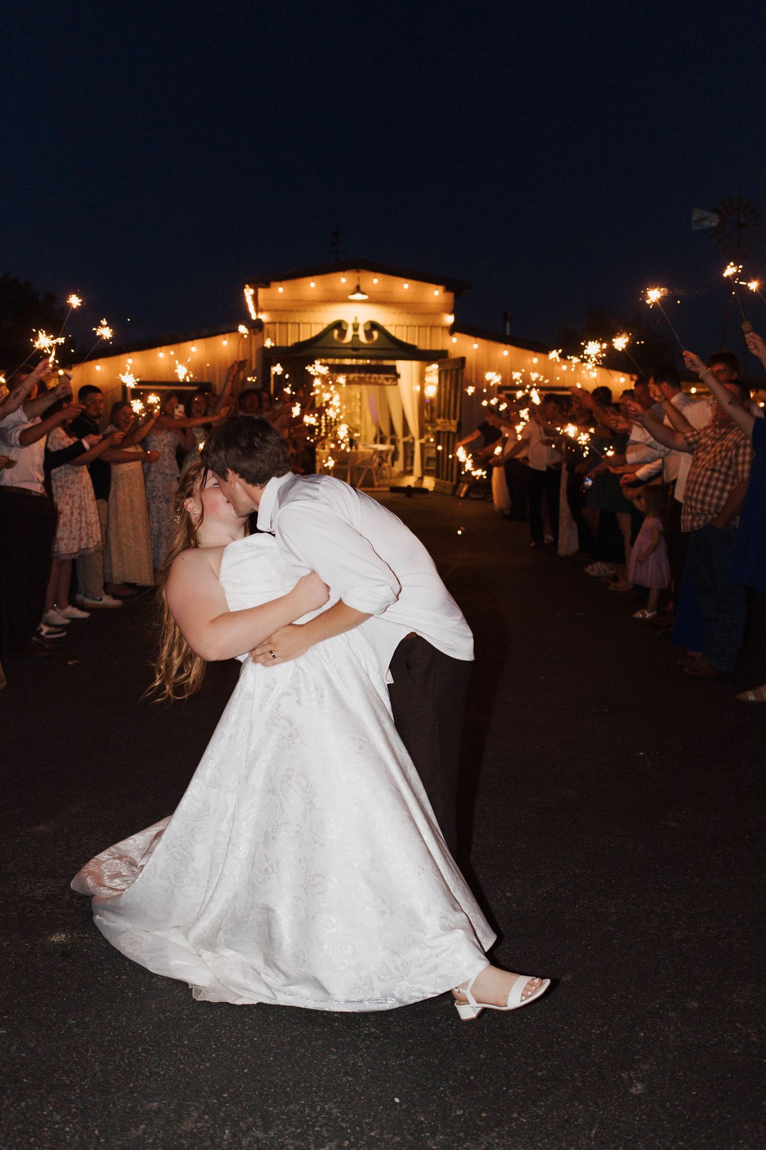 Newlyweds share a romantic dip kiss during their sparkler sendoff at a warmly lit wedding venue at night.