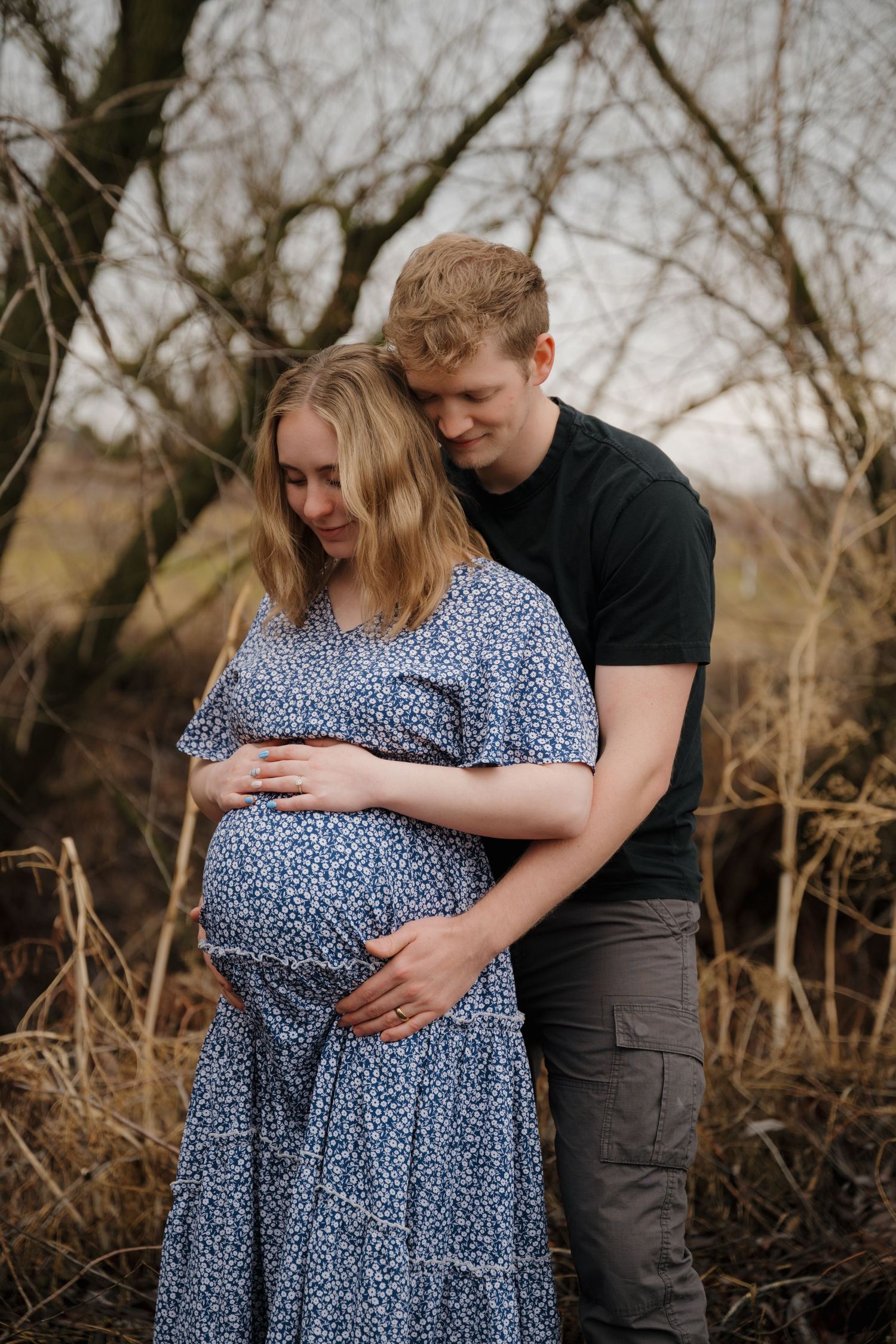 Tender maternity moment captured outdoors with a couple embracing among bare winter trees.