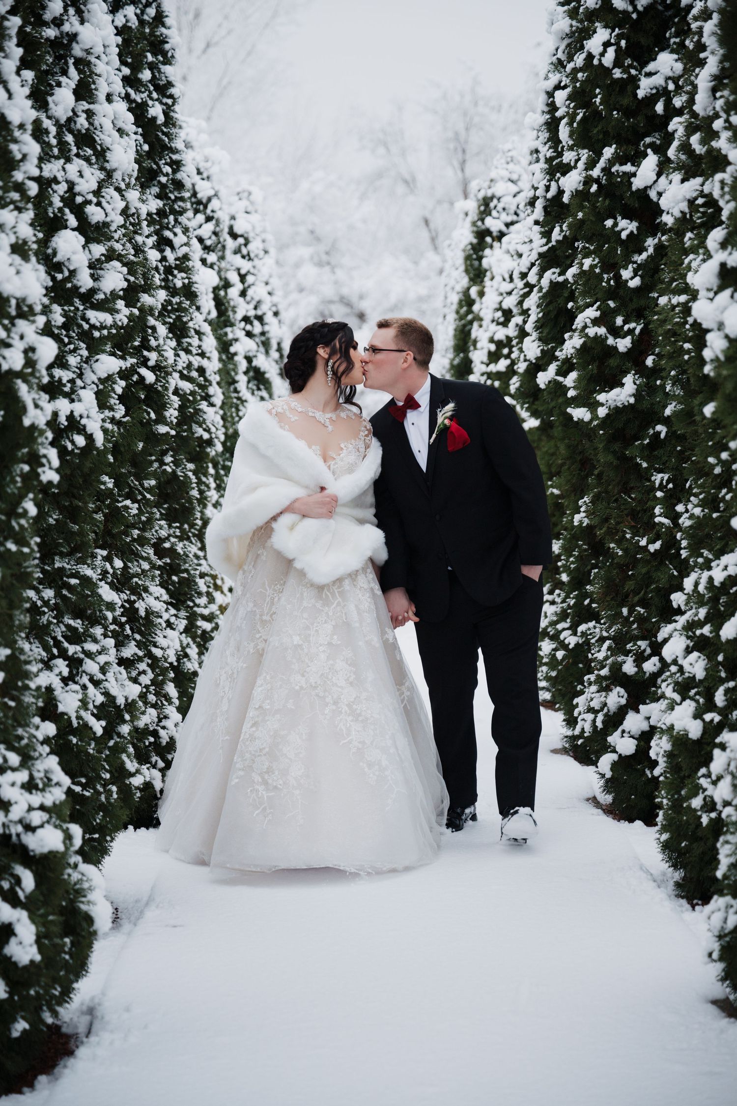 A romantic winter wedding photo with snow-covered evergreens creating a magical garden pathway corridor.