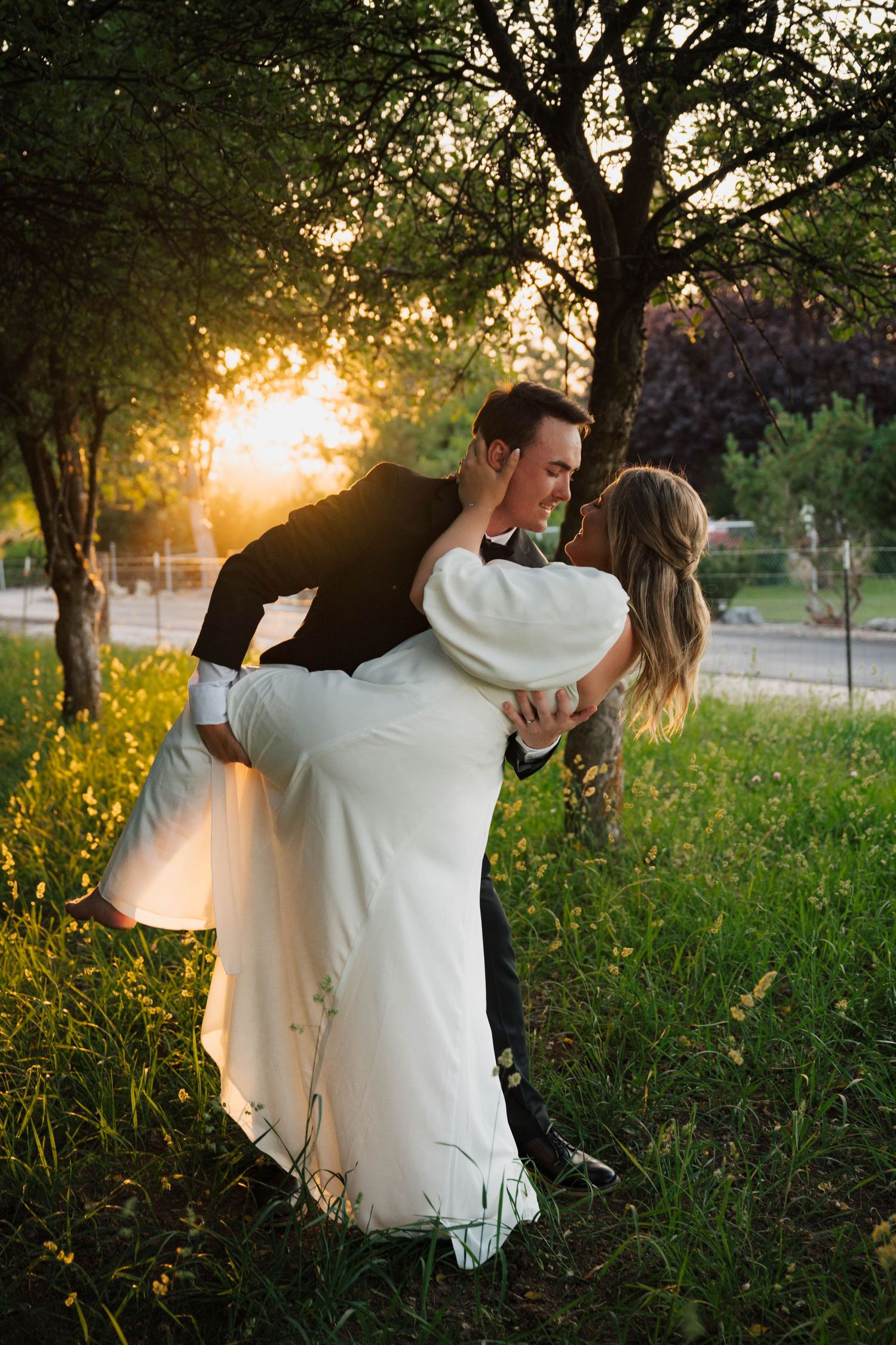 A romantic couple shares a dip kiss at sunset in a grassy park with golden sunlight filtering through trees.