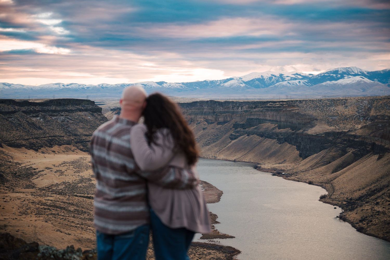A couple embraces while overlooking a scenic river canyon with snow-capped mountains in the distance.