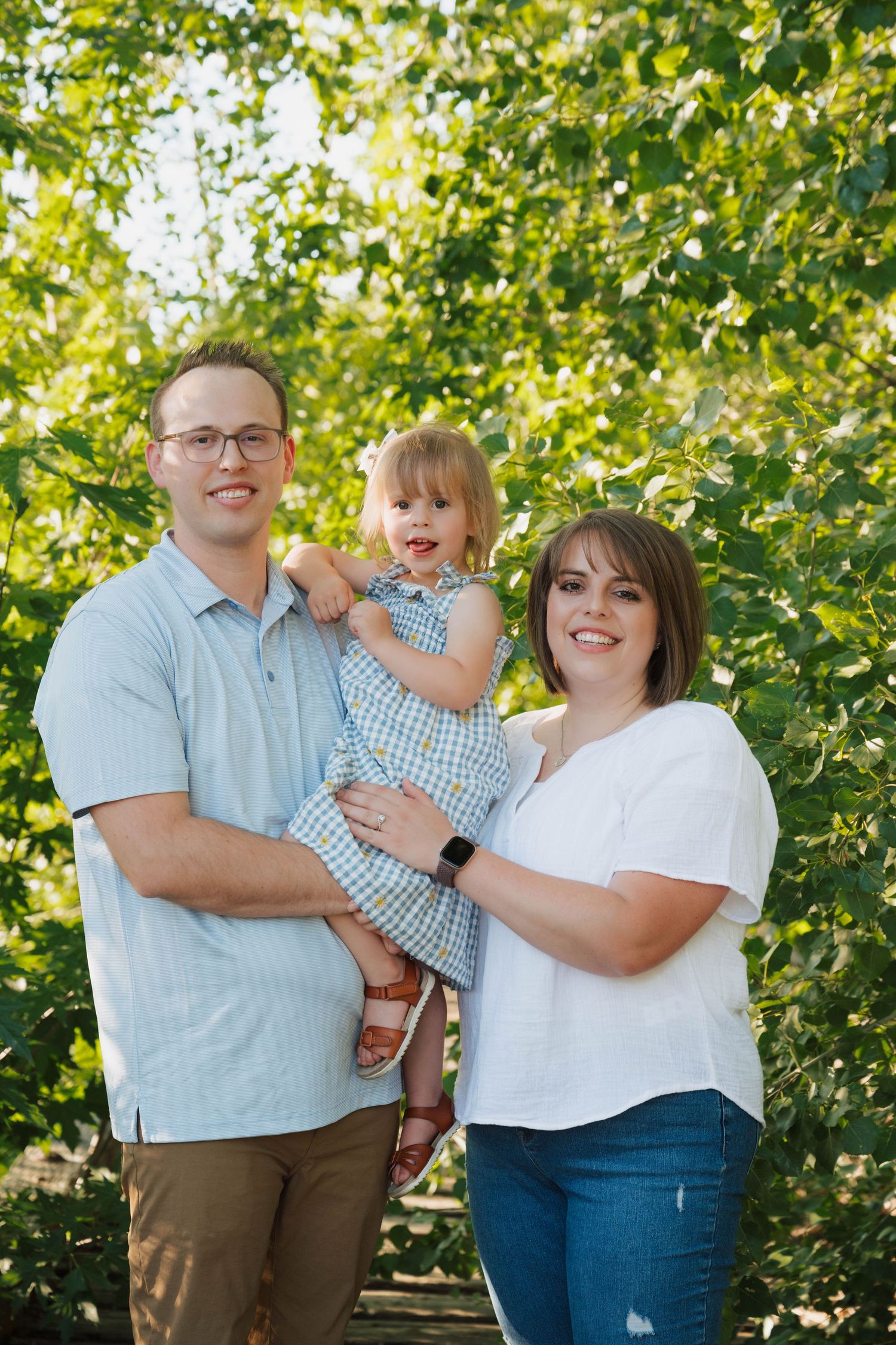 A smiling family poses together outdoors against green leafy trees in summer clothing.