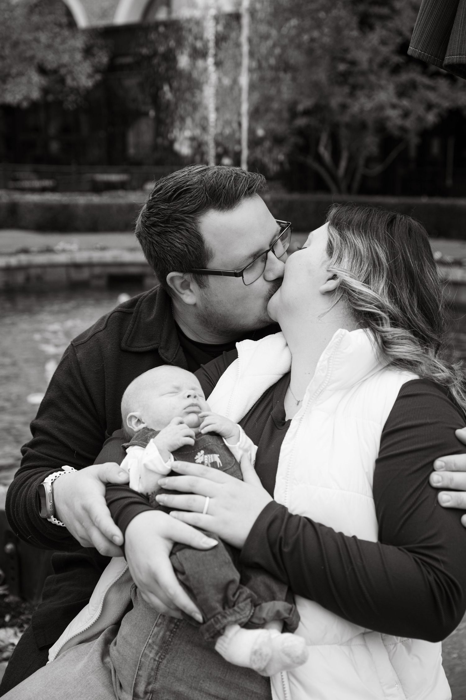 Parents share an intimate moment while holding their newborn baby outdoors.