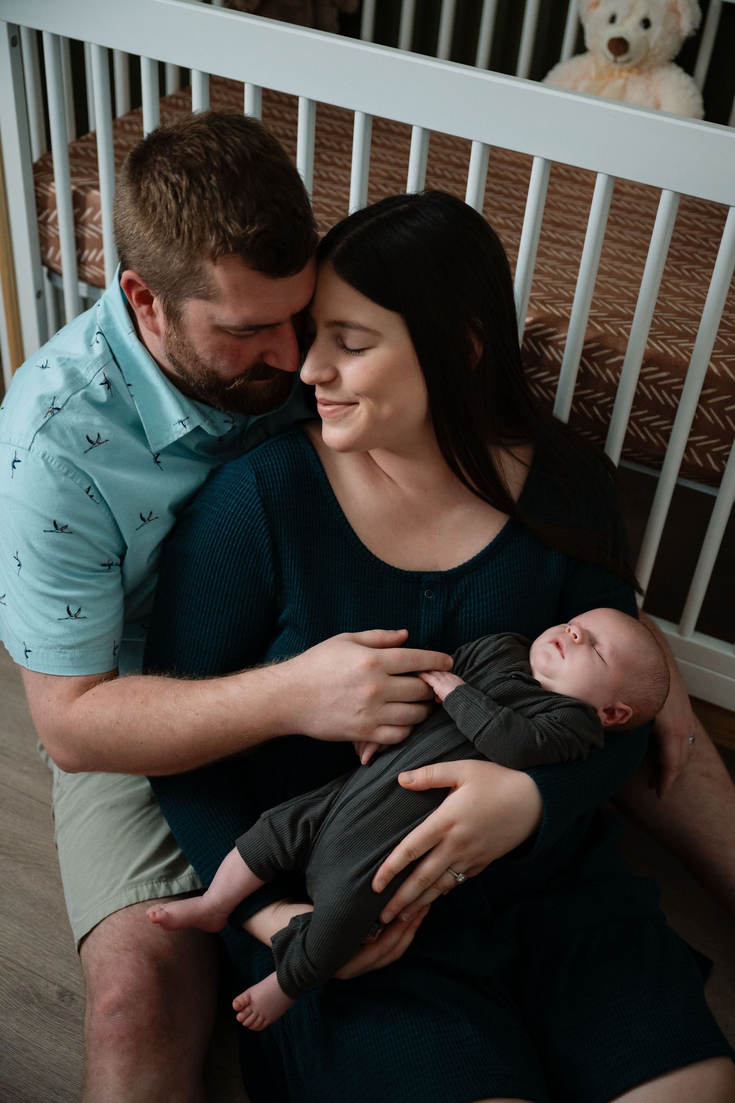 Family shares an intimate moment with their newborn baby in front of a white crib with a teddy bear.