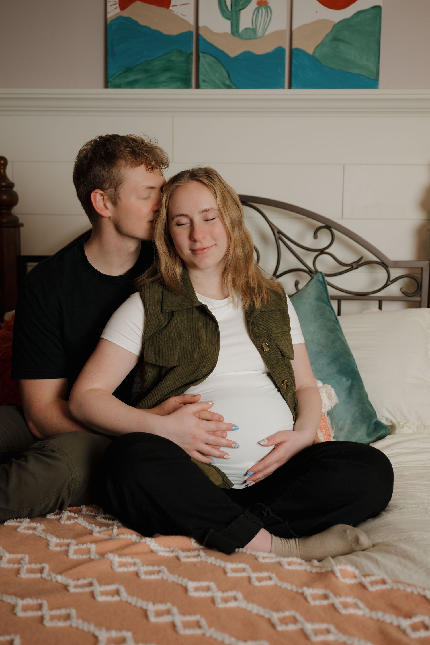 Intimate maternity portrait with couple sitting on bed beneath colorful wall art.
