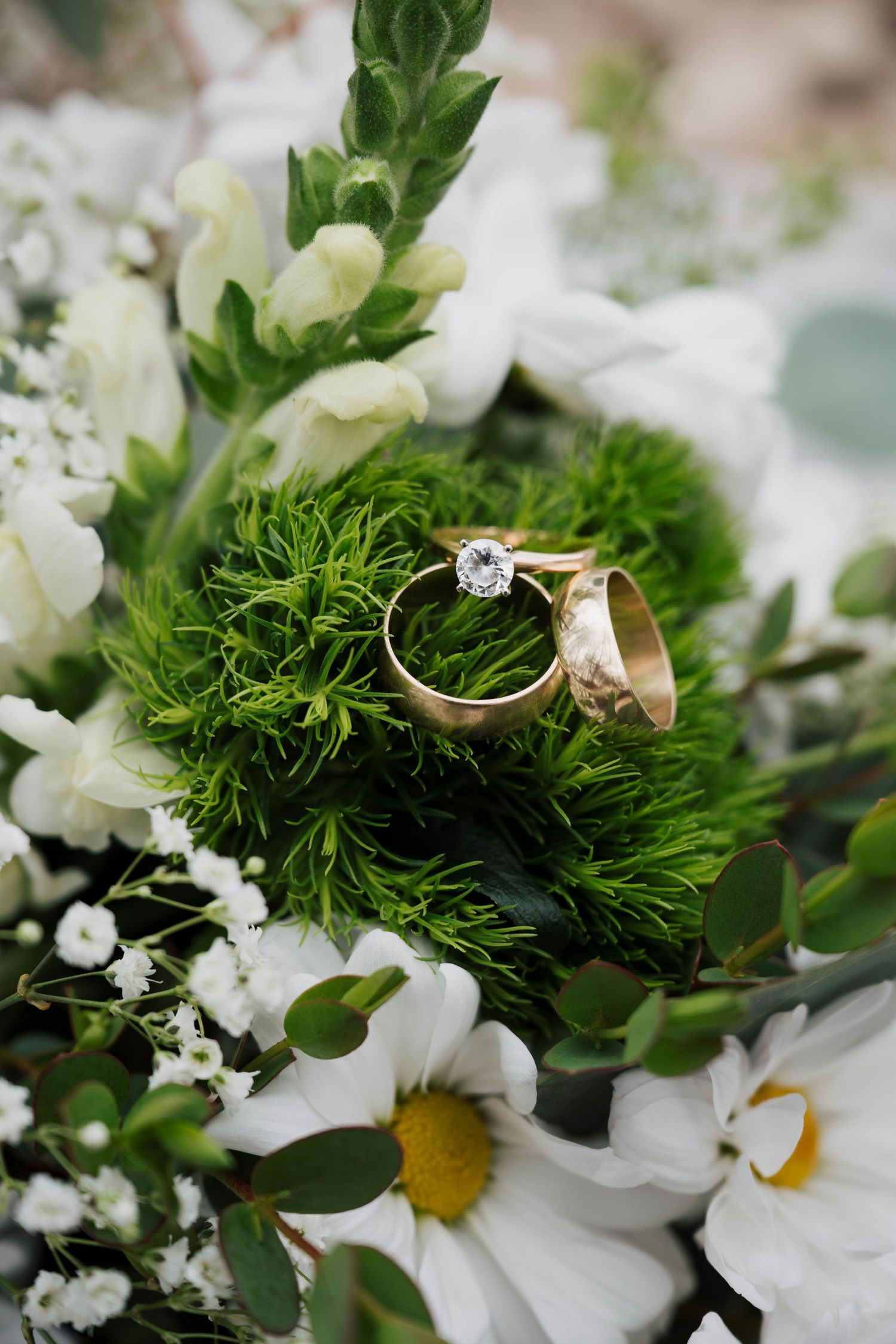 Gold wedding bands and diamond engagement ring resting on green moss with white daisies and floral decor.