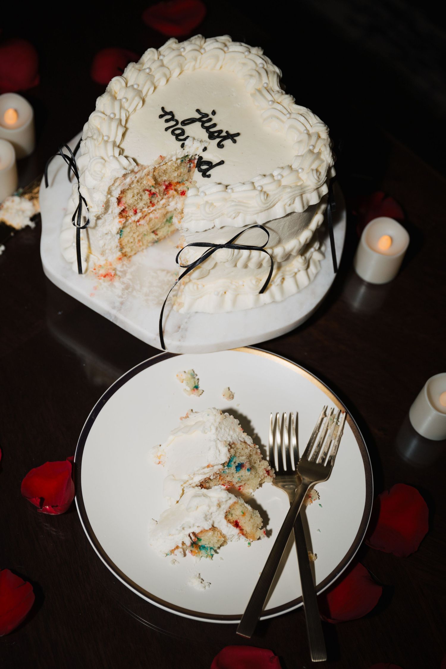 Heart-shaped white wedding cake with a slice cut out, displayed with candles and rose petals.