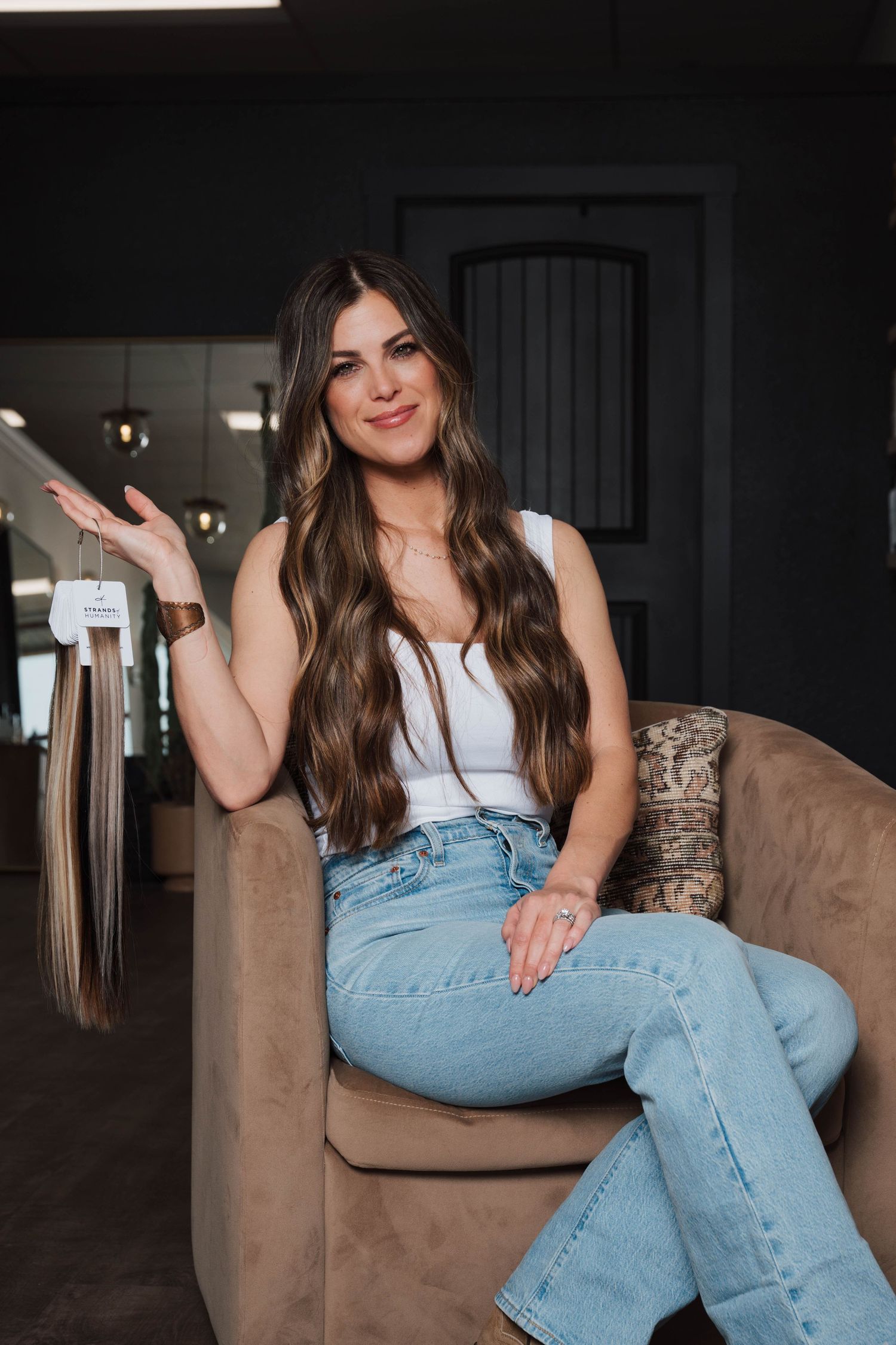 A casual lifestyle portrait showing someone in a white tank top and light wash jeans sitting on a brown armchair.