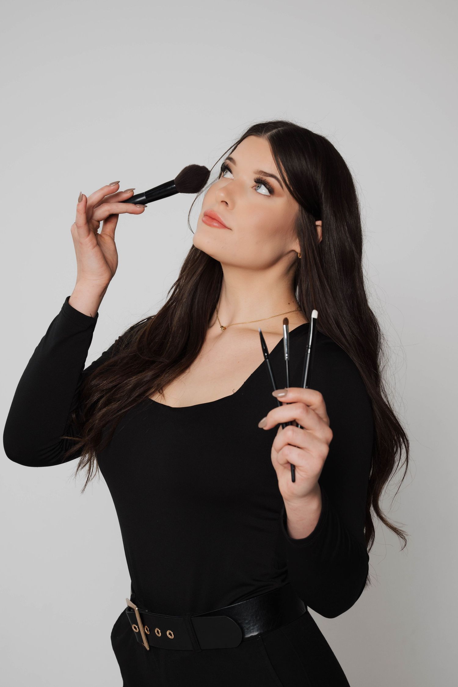 A makeup artist in a black outfit holds brushes and tools while applying makeup, looking upward against a light background.
