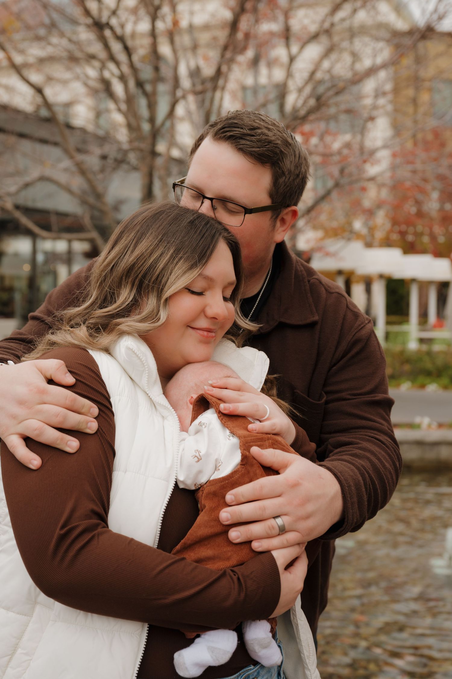 Family embraces while holding their newborn baby near a waterfront in autumn.